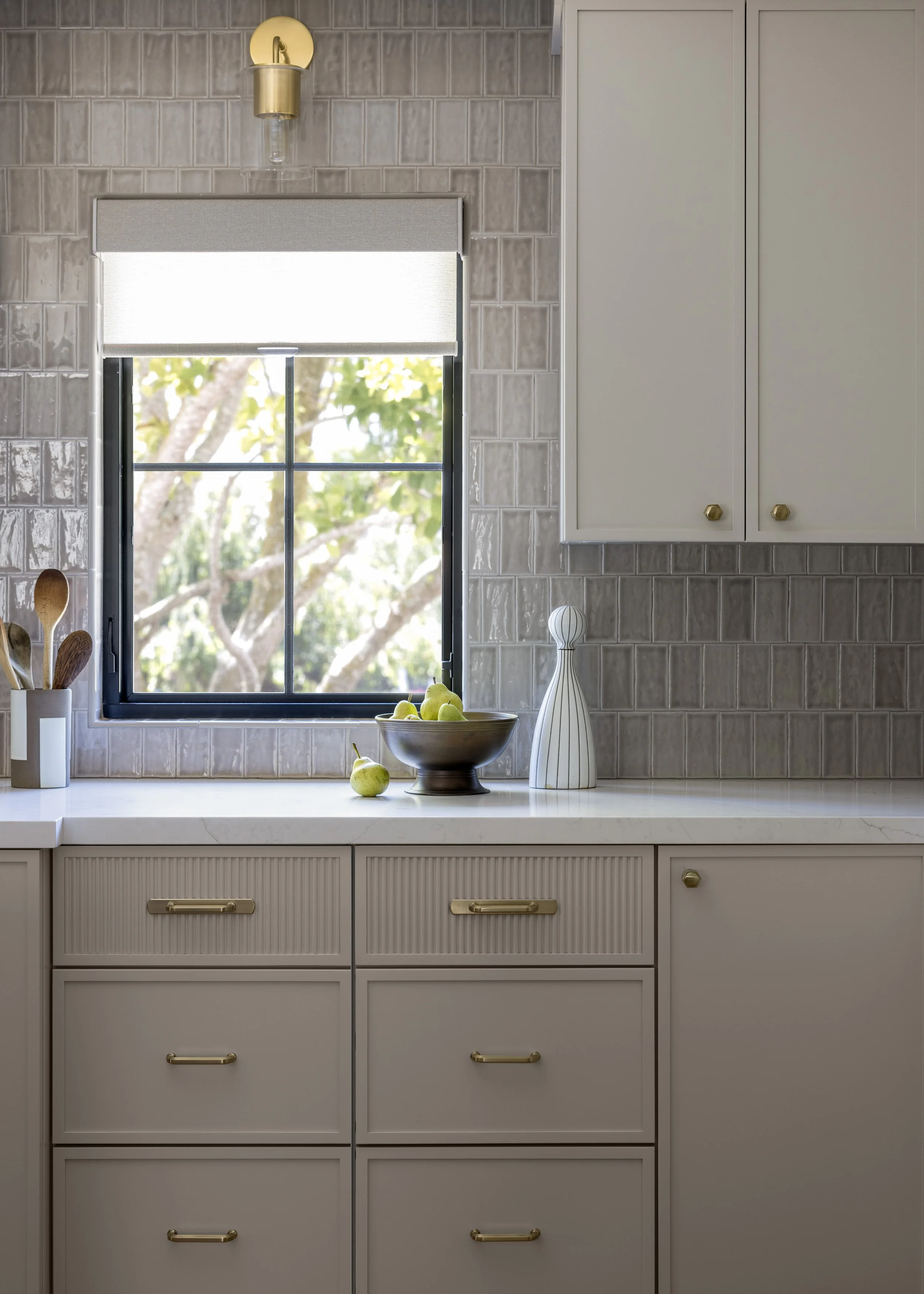 Kitchen counter with a bowl of pears, a tall white vase, and a white cabinet with gold knobs, window with a view of trees outside, and a wall-mounted light fixture.