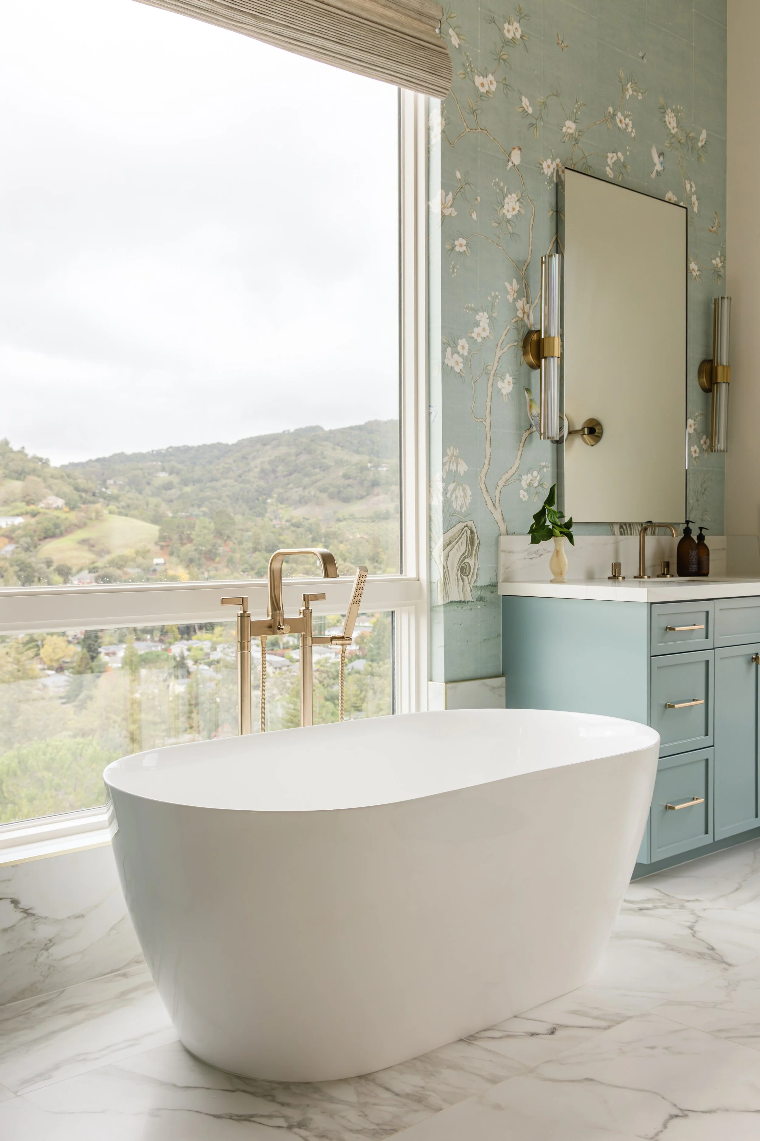 Modern bathroom with a freestanding oval bathtub, marble flooring, a large window with a view of green hills, blue cabinetry, and a mirror with wall-mounted lights.
