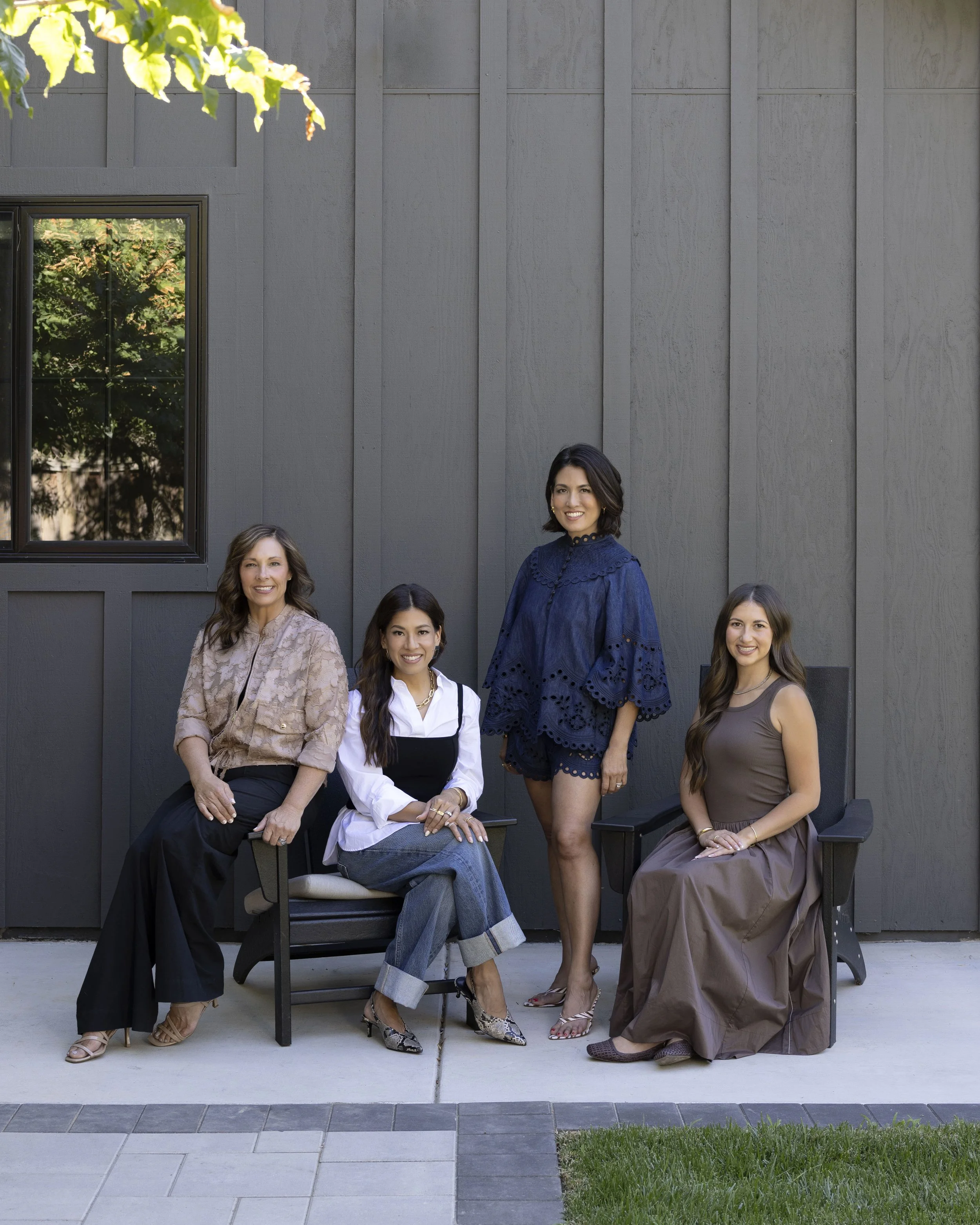 Five women posing outdoors in front of a gray wooden wall, some sitting and others standing, dressed in stylish clothing and smiling.