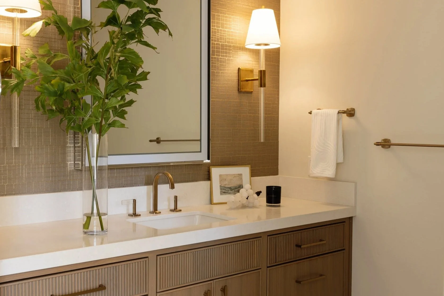 A modern bathroom vanity with a white countertop, a gold faucet, and a large mirror. On the countertop, there is a tall glass vase with green leaves, a framed picture, a cluster of white decorative balls, and a black speaker. The background includes a textured brown wall with a wall-mounted lamp, a white towel on a gold towel bar, and a walk-in shower with glass enclosure and gold fixtures.