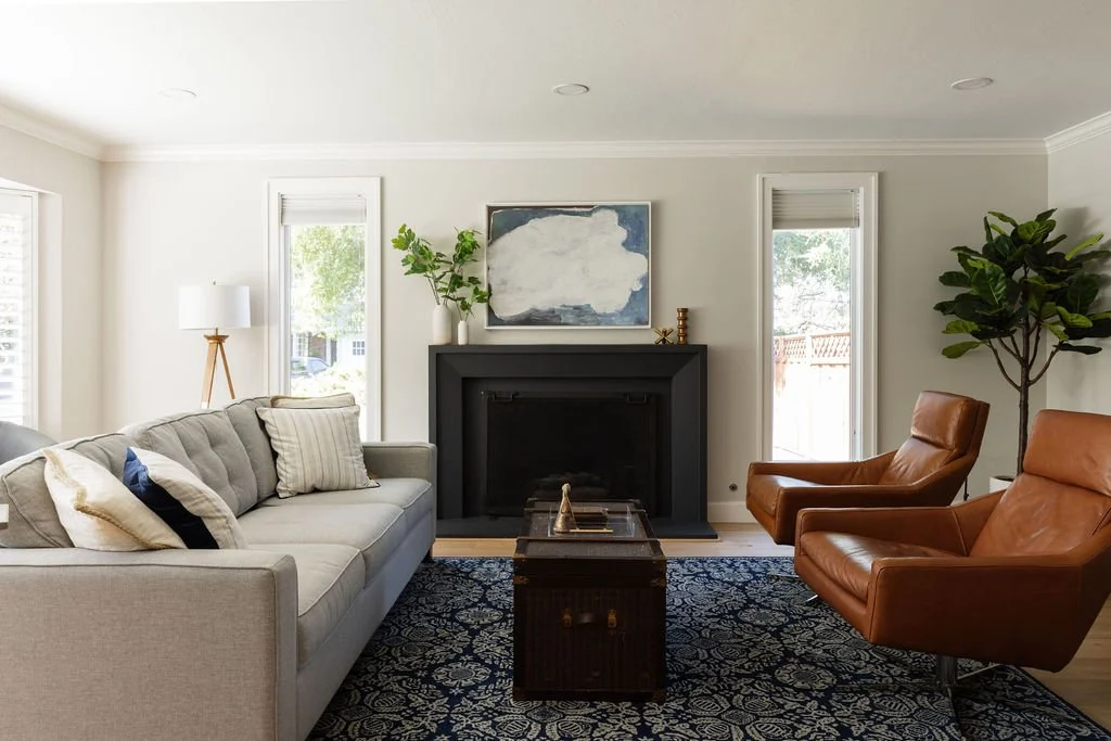Living room with a light-colored sofa, two brown leather chairs, a black fireplace with artwork above, a geometric patterned area rug, and a large plant to the right.