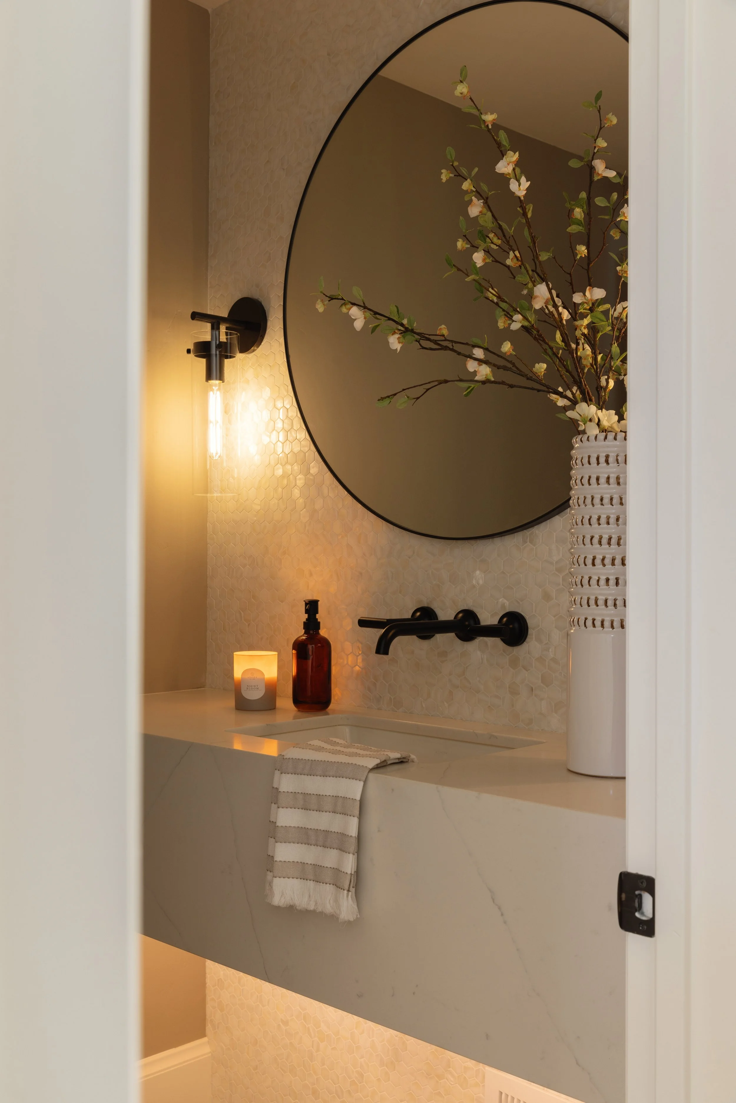 A cozy bathroom with a large round mirror, a black wall-mounted light, a vase with blooming branches, a lit candle, and a brown soap dispenser on a marble countertop. There is a striped towel hanging over the edge of the sink.