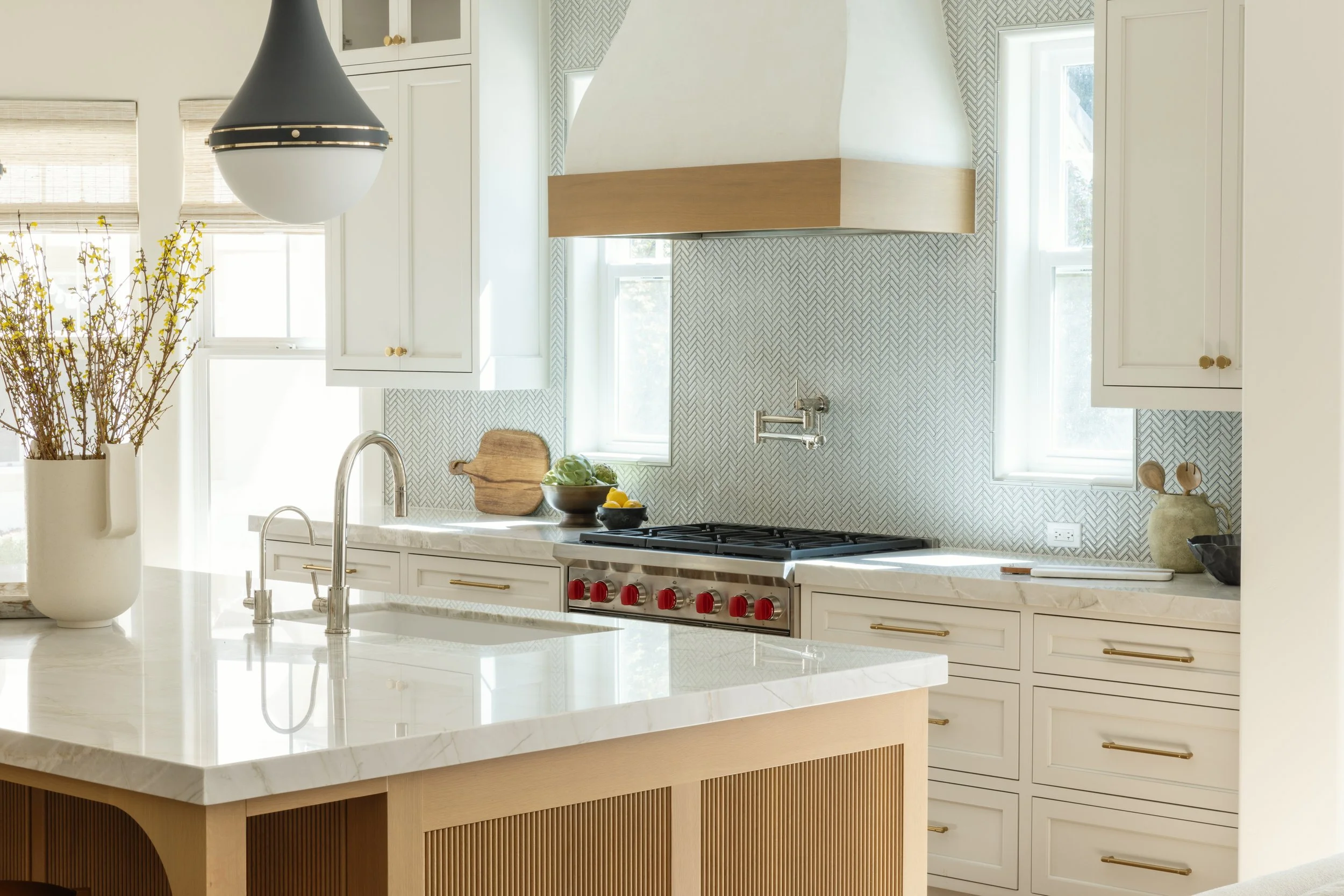 A modern kitchen with white cabinetry, a marble island with a wooden base, a black and white range hood, a stainless steel faucet, a vase with yellow branches, and two windows letting in natural light.