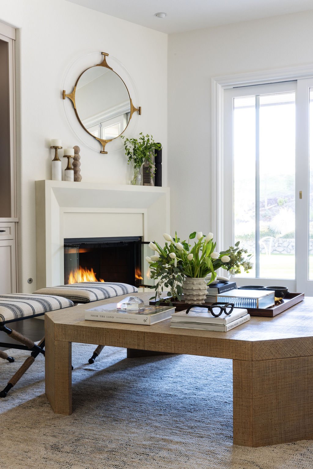 Living room with white fireplace, large window, wooden coffee table with a vase of white flowers, books, glasses, and box on it. Blue and white bench, wall mirror, candles, and decorative objects on the mantle.