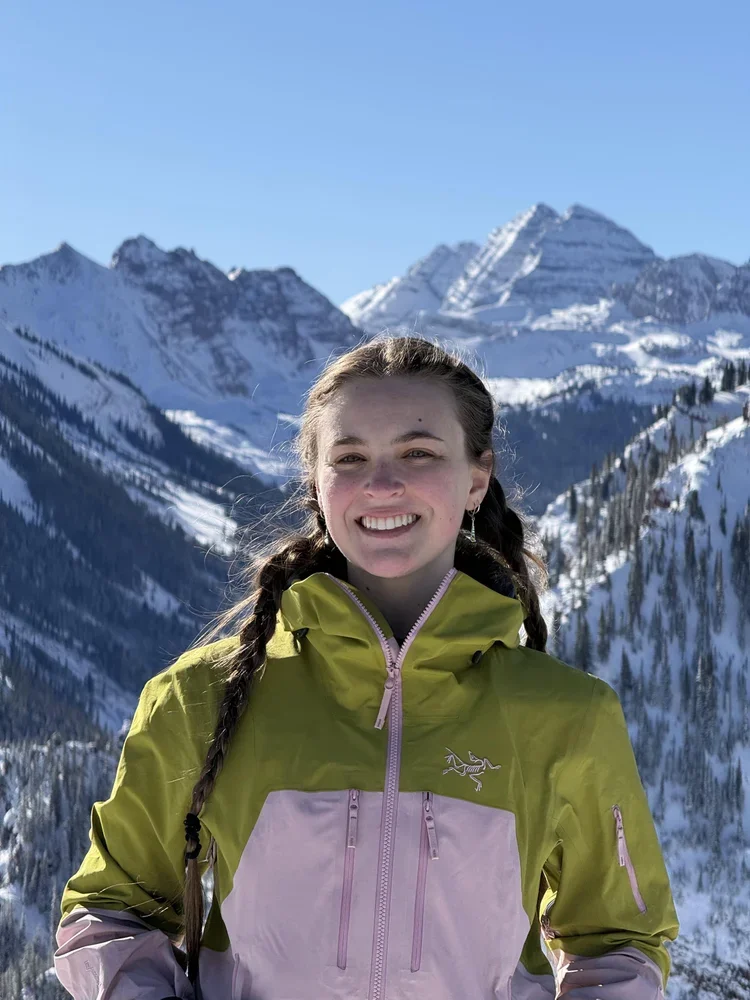 A young woman with braided hair smiling in front of snowy mountains under a clear blue sky, wearing a yellow and pink outdoor jacket.