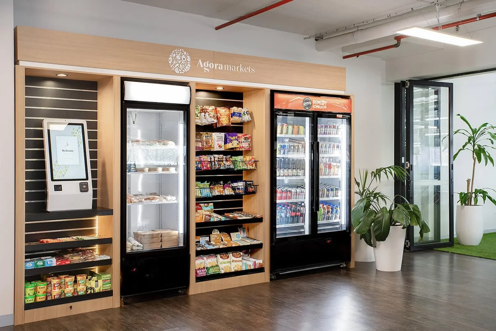 A modern vending area with three refrigerated vending machines and a self-checkout kiosk, decorated with potted plants.