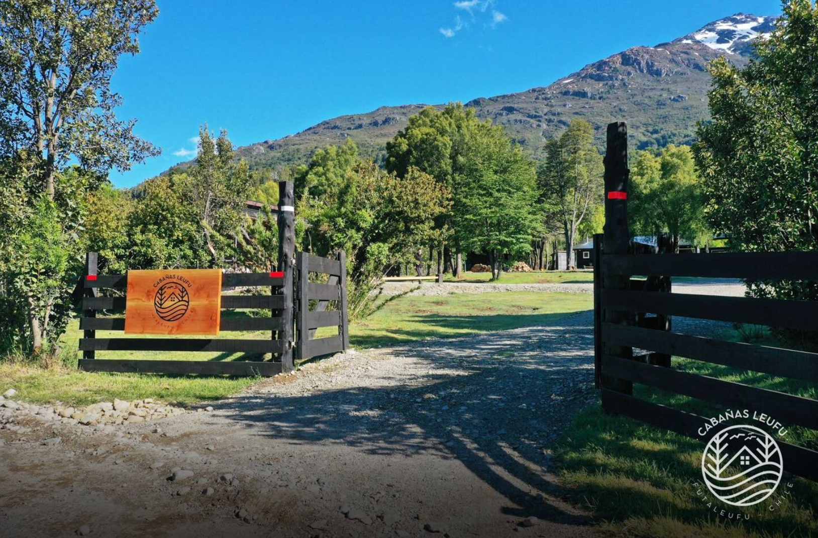 Vista de una entrada a una cabaña rodeada de árboles, con montañas en el fondo y un cielo azul despejado. Hay puertas de madera de color negro, una de ellas con un logo de círculo que tiene una casa, árboles y un río, y un cartel naranja que dice "Ca