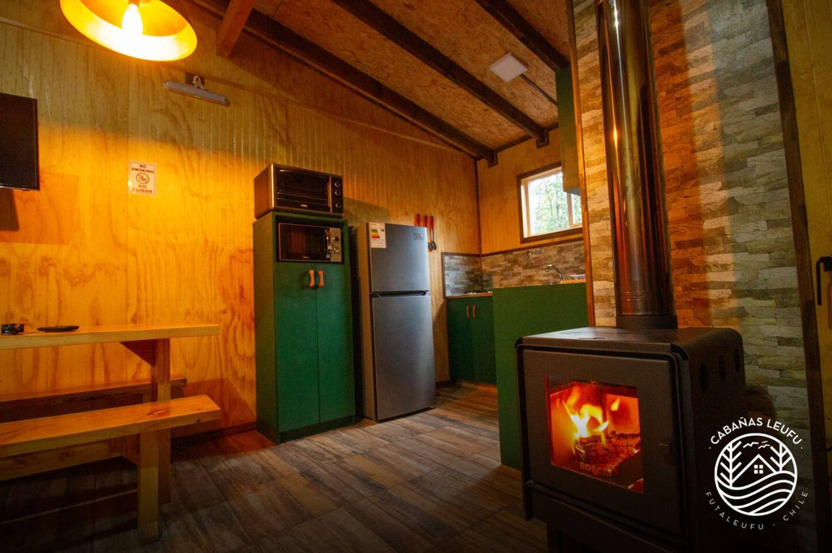Interior de una cabaña de madera con cocina equipada, una estufa de leña encendida, refrigerador, armario verde, mesa de madera y ventanas que dejan entrar luz natural.