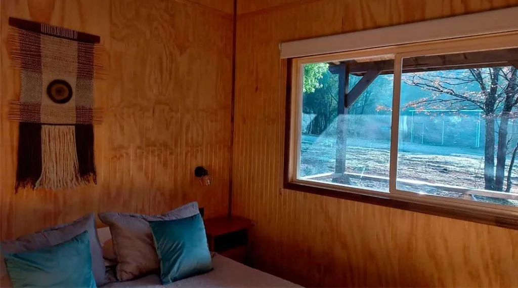 Habitación con paredes de madera, ventana grande que muestra árboles y césped exterior, cama con almohadas color azul y gris, decoraciones en la pared.