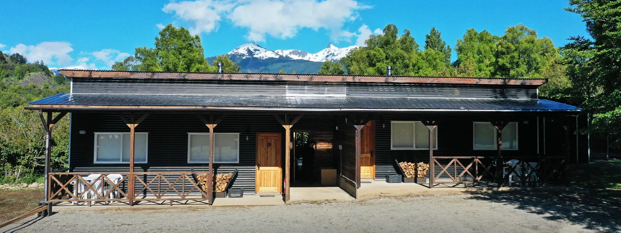 Casa de madera con balcón y leña apilada en un lado, rodeada de árboles y montañas con nieve en el fondo, cielo azul con nubes.