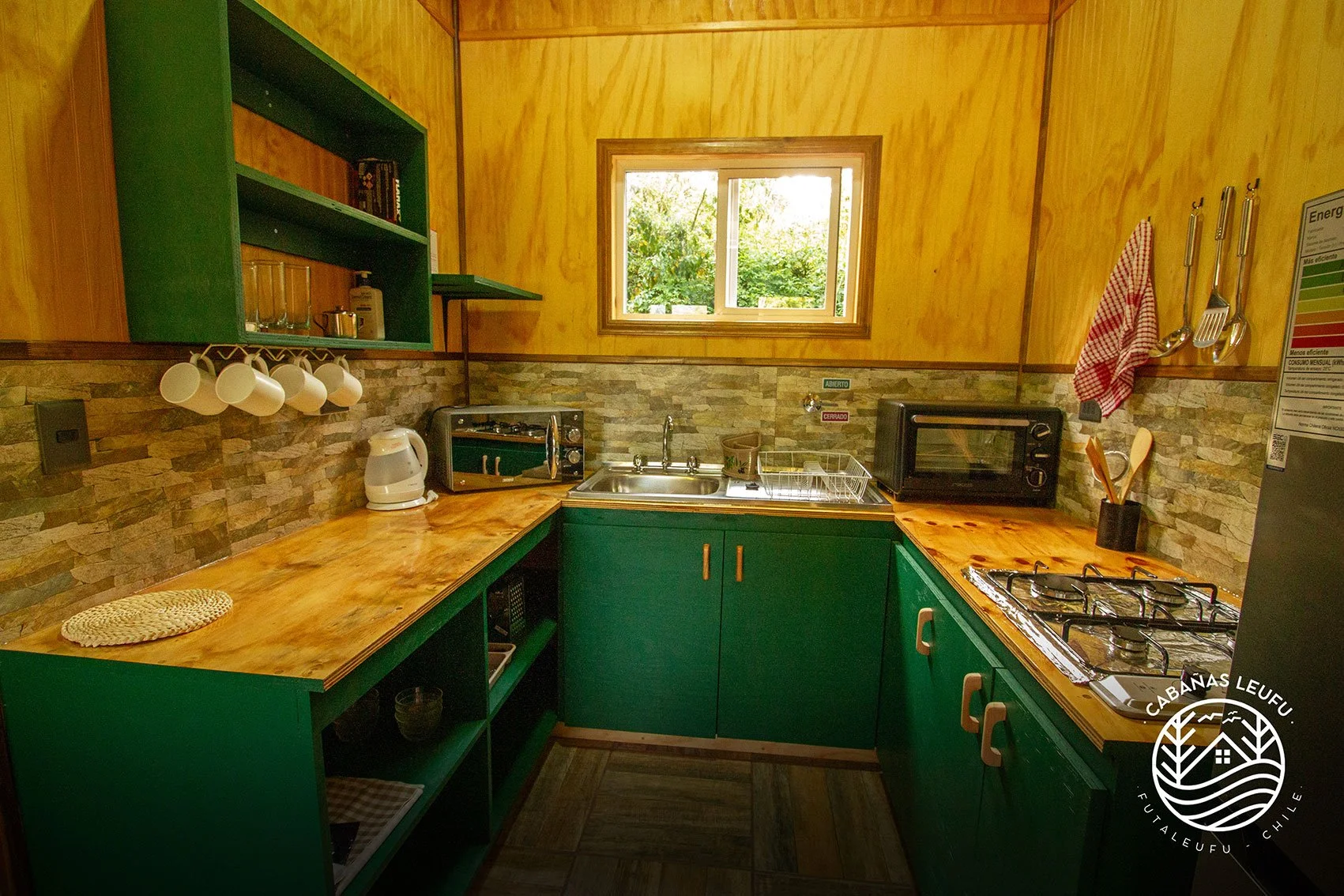 Cocina de cabaña con paredes de madera, encimeras de madera, gabinetes verdes, estantes con utensilios y tazas, y un horno microondas. Ventana pequeña con vista a árboles y naturaleza.