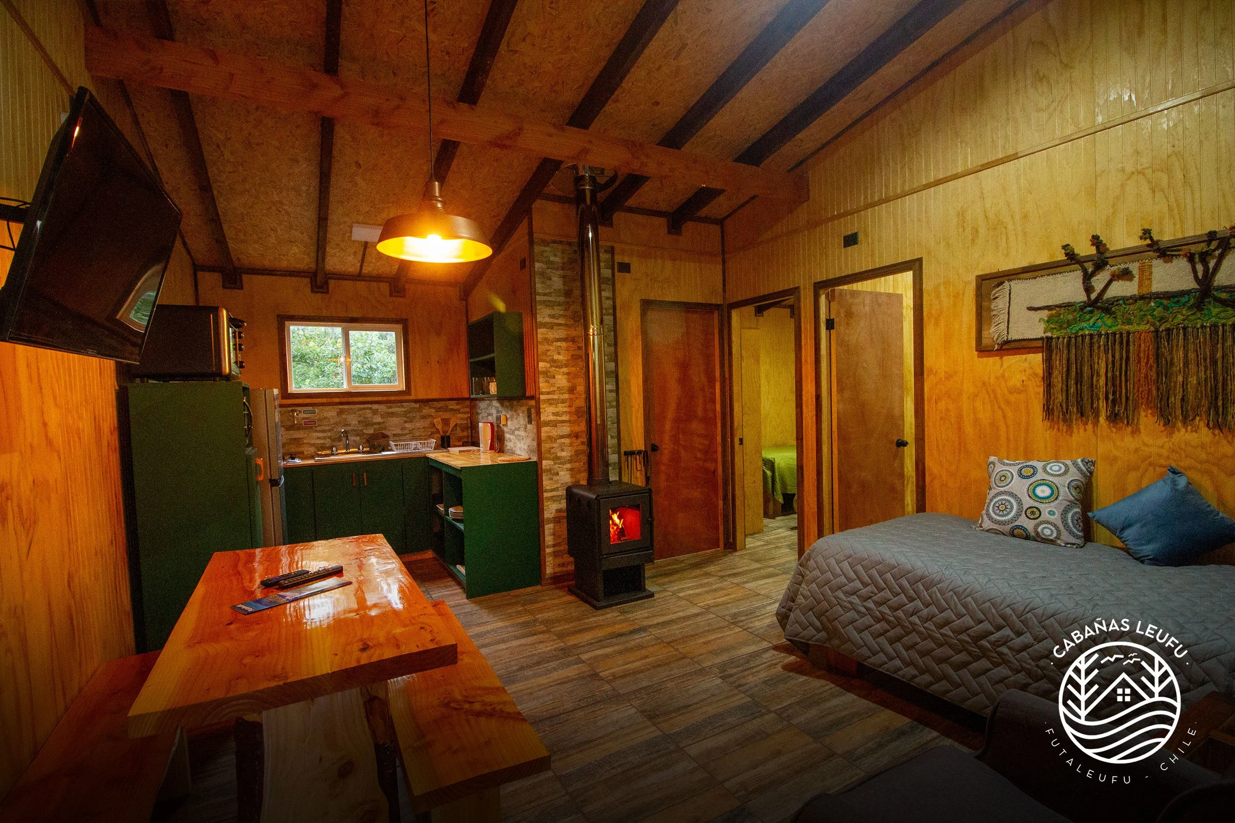 Interior de una cabaña de madera con sala y cocina, con una cama con almohadas, una estufa de leña, paredes de madera, piso de madera y una lámpara colgante. Ventana al fondo y puerta a habitaciones.