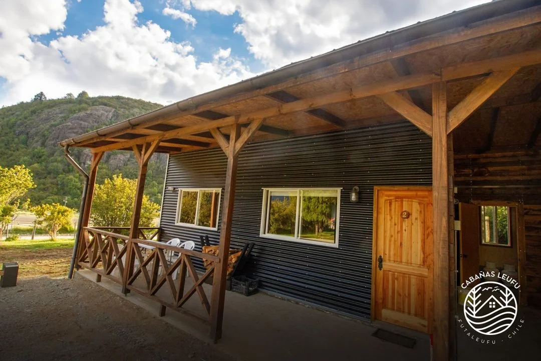 Vista frontal de una cabaña con porch de madera, en un entorno natural con árboles y montañas, cielo nublado.