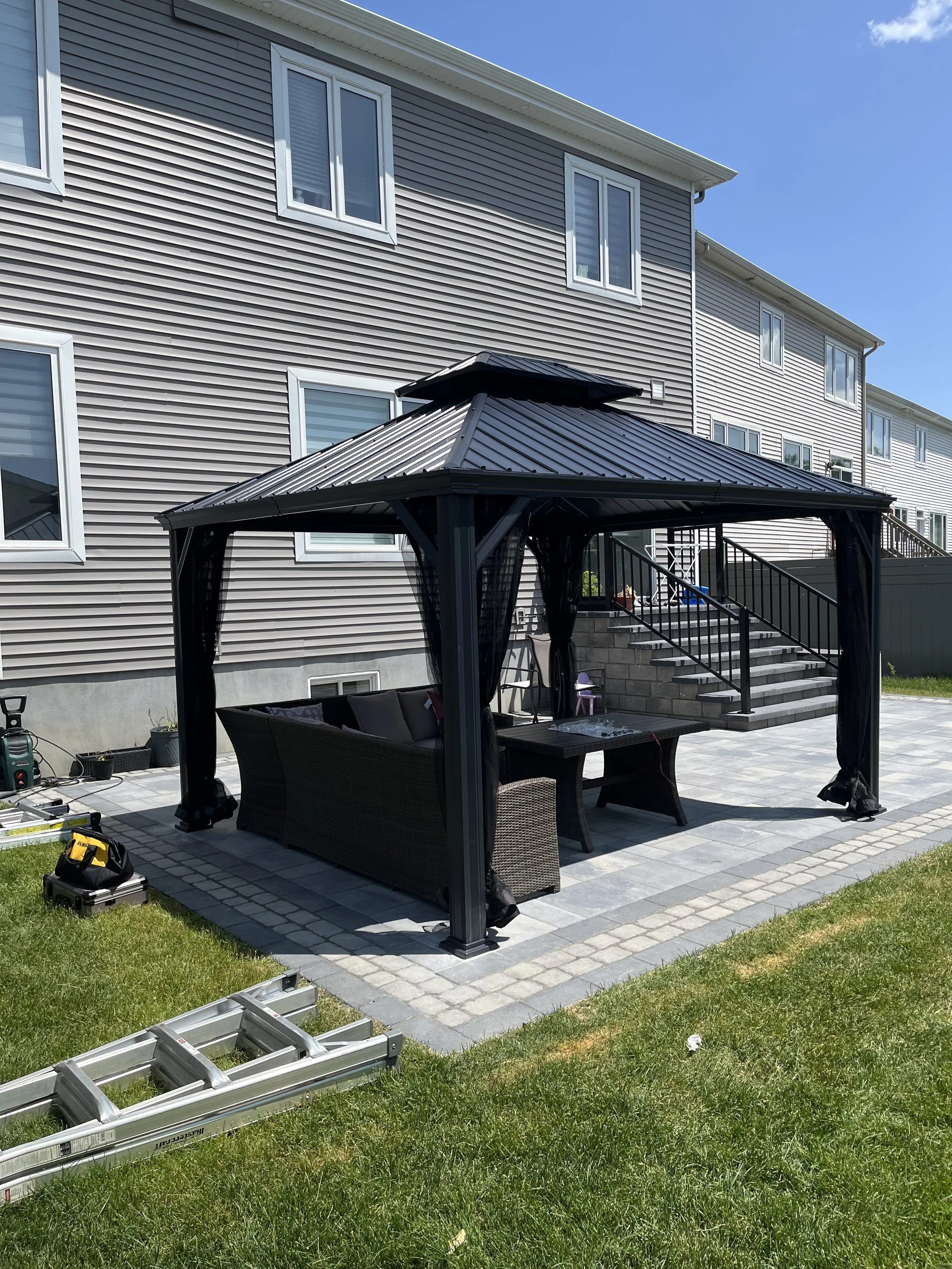 A backyard patio with a black gazebo, wicker patio furniture with cushions, and a small table. There are stairs leading up to a house with gray siding. Construction tools and a ladder are on the grass nearby.