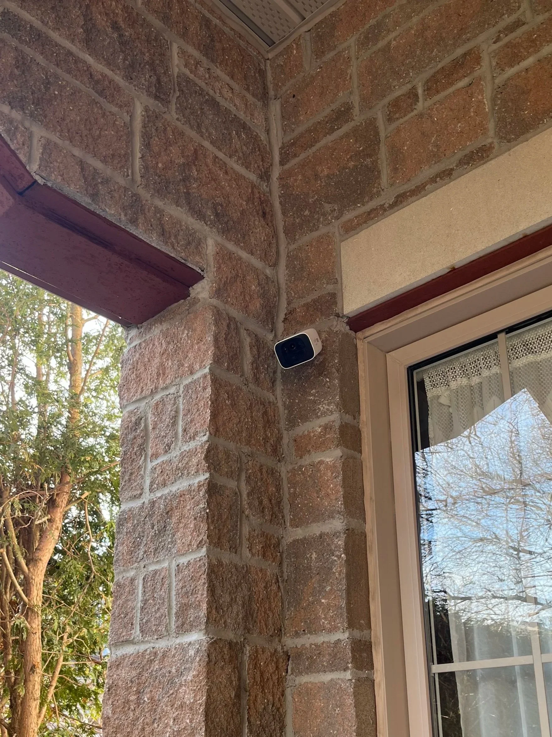 Close-up of an exterior house corner with brick and stucco wall, a security camera mounted near the window, and a tree visible outside.