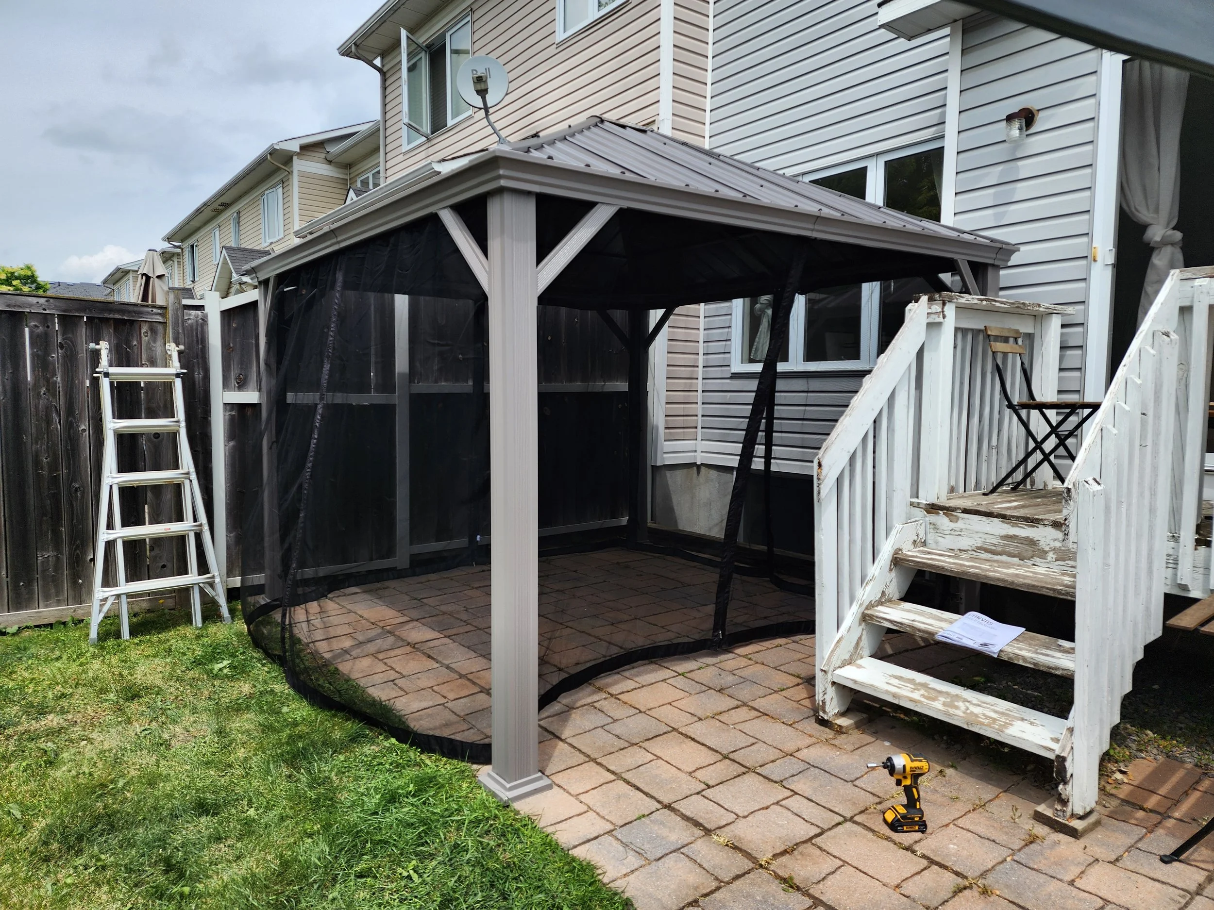 Backyard patio with a partially assembled screened gazebo, a wooden staircase leading to a house, a step ladder, a cordless drill, and a small folding chair.
