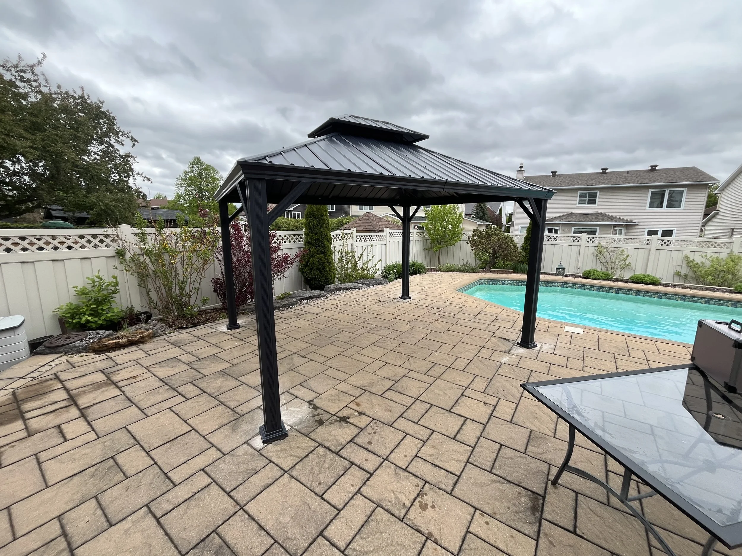 Backyard with a rectangular swimming pool, a black metal gazebo, paver patio, and a white fence with neighboring houses in the background.