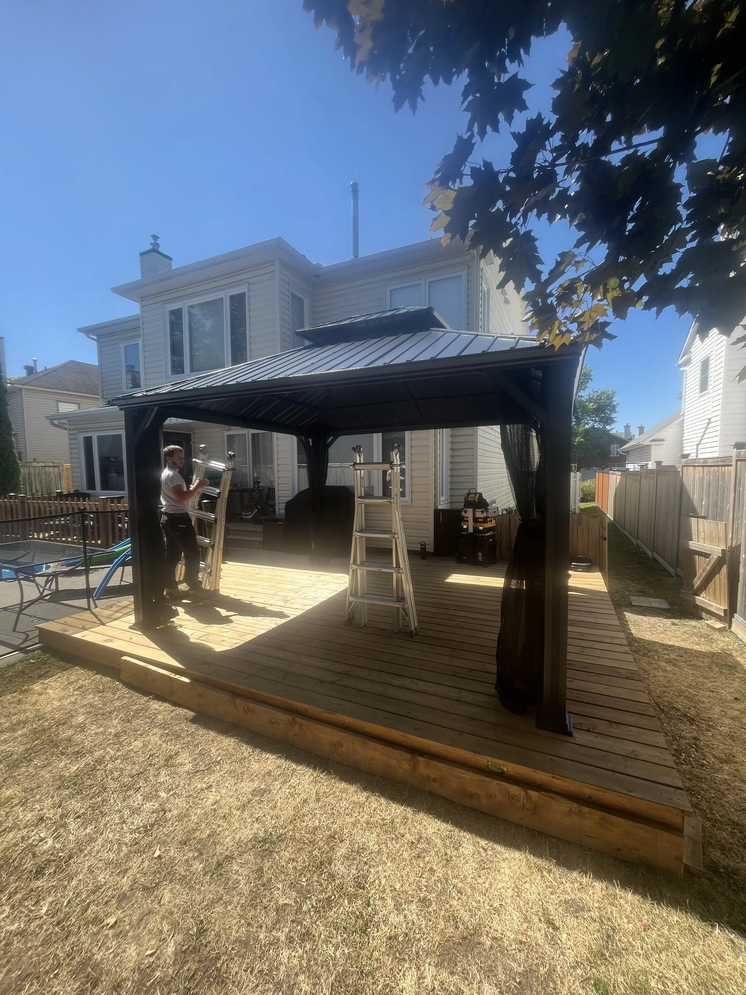 A person standing on a wooden deck in a backyard, working with ladders under a black metal-roofed gazebo, with a white house in the background and a clear blue sky.