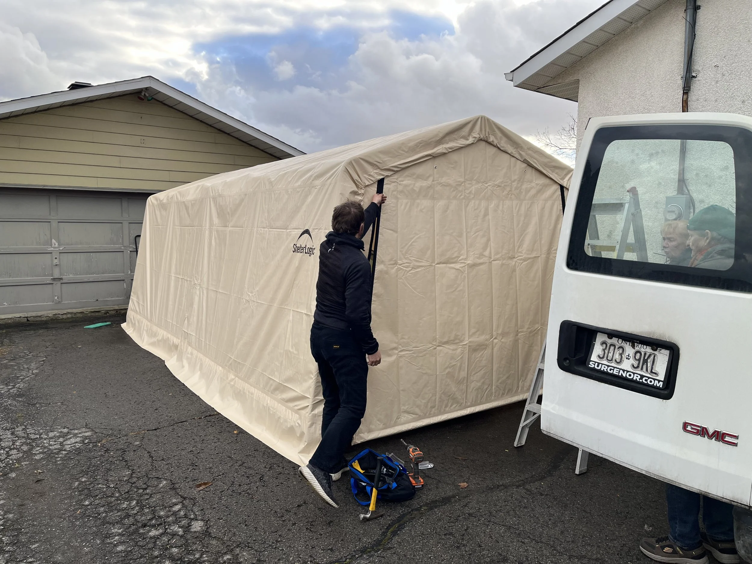 Two men setting up a beige outdoor tent next to a white GMC van, with a yellow house and cloudy sky in the background.
