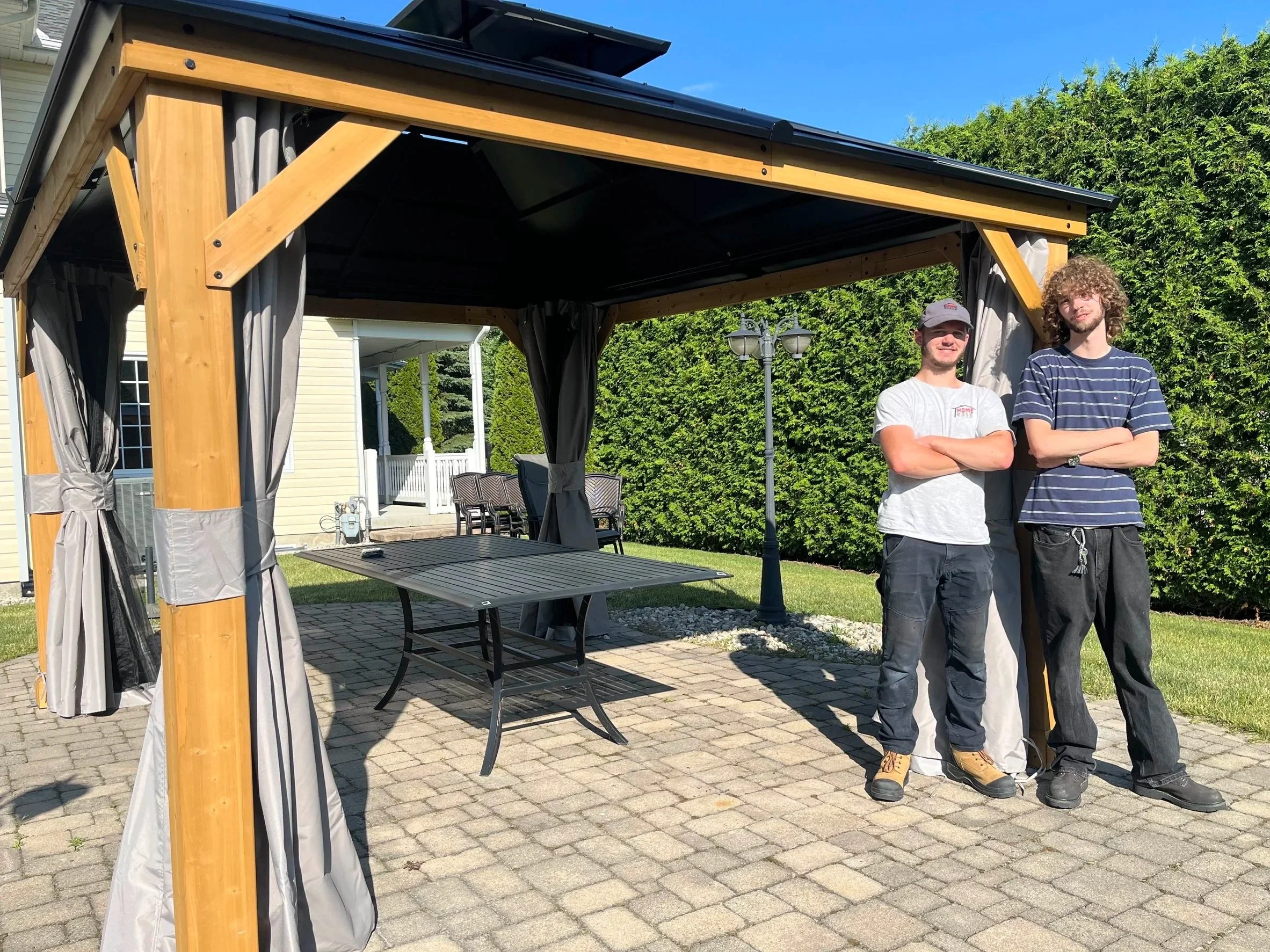 Two young men standing under a wooden gazebo outdoors on a sunny day, with a house, green bushes, a brick patio, and a lamp post in the background.