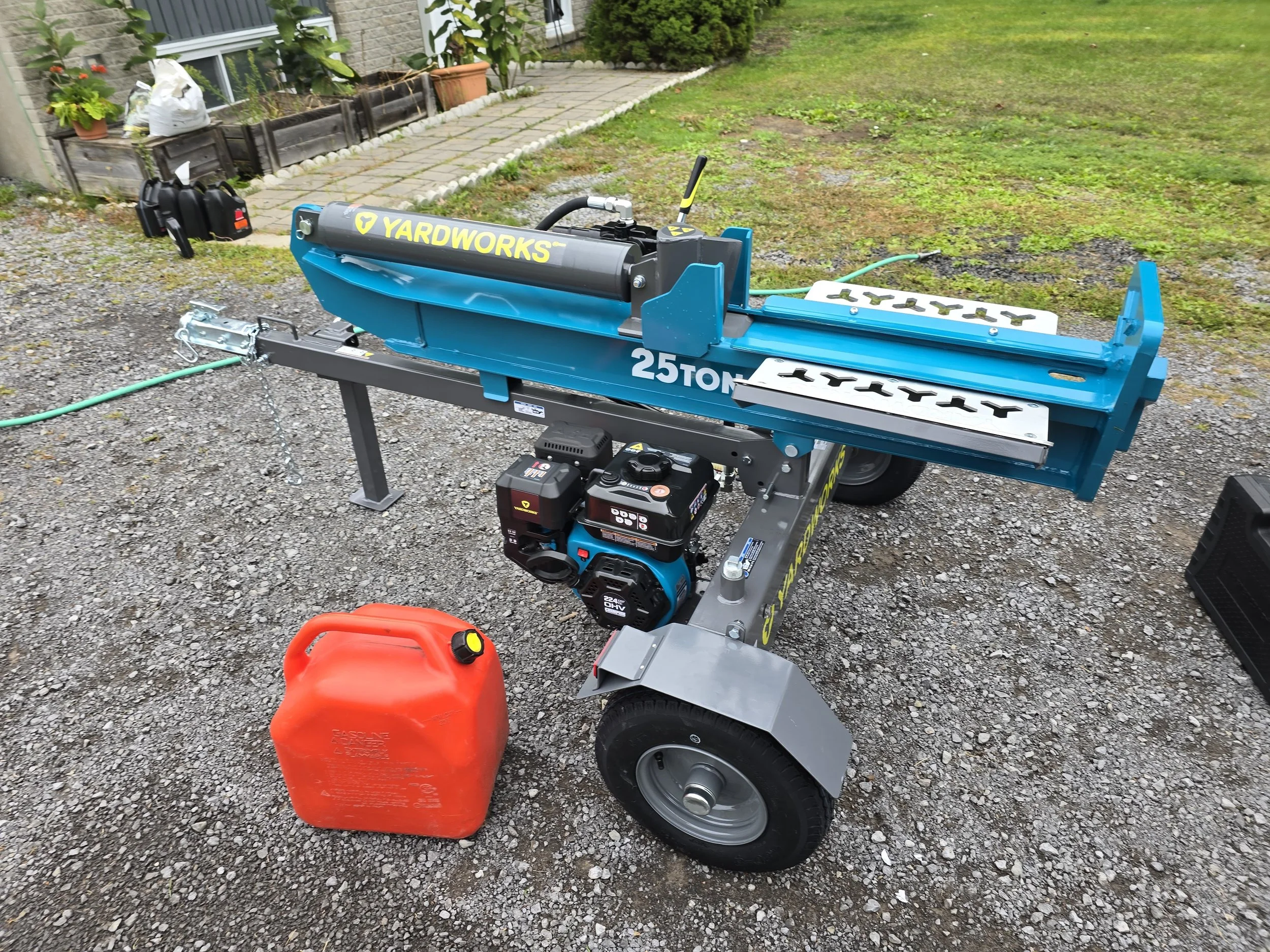 A portable, blue Yardworks 25-ton log splitter with a gasoline engine, set on a gravel surface outdoors. A red fuel container is nearby, and a garden is visible in the background.