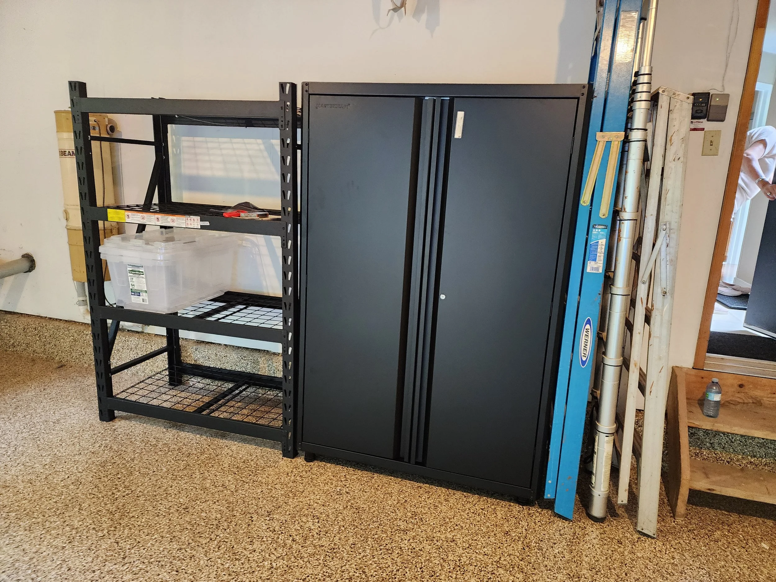 Black metal shelving unit with some tools and a plastic container, next to a black storage cabinet, installed against a white wall in a garage or workshop.