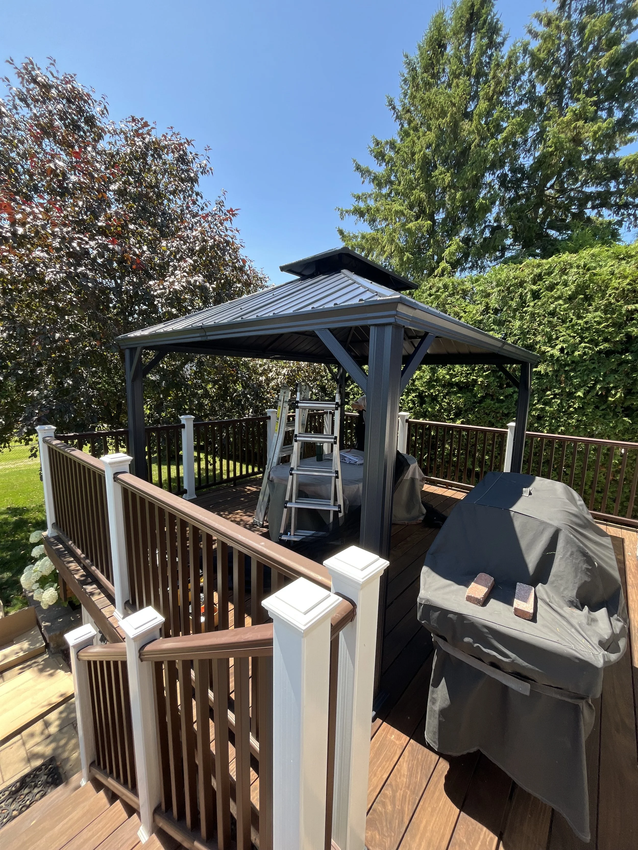 A backyard deck with a gazebo, a ladder, and a covered grill, surrounded by green trees and blue sky.