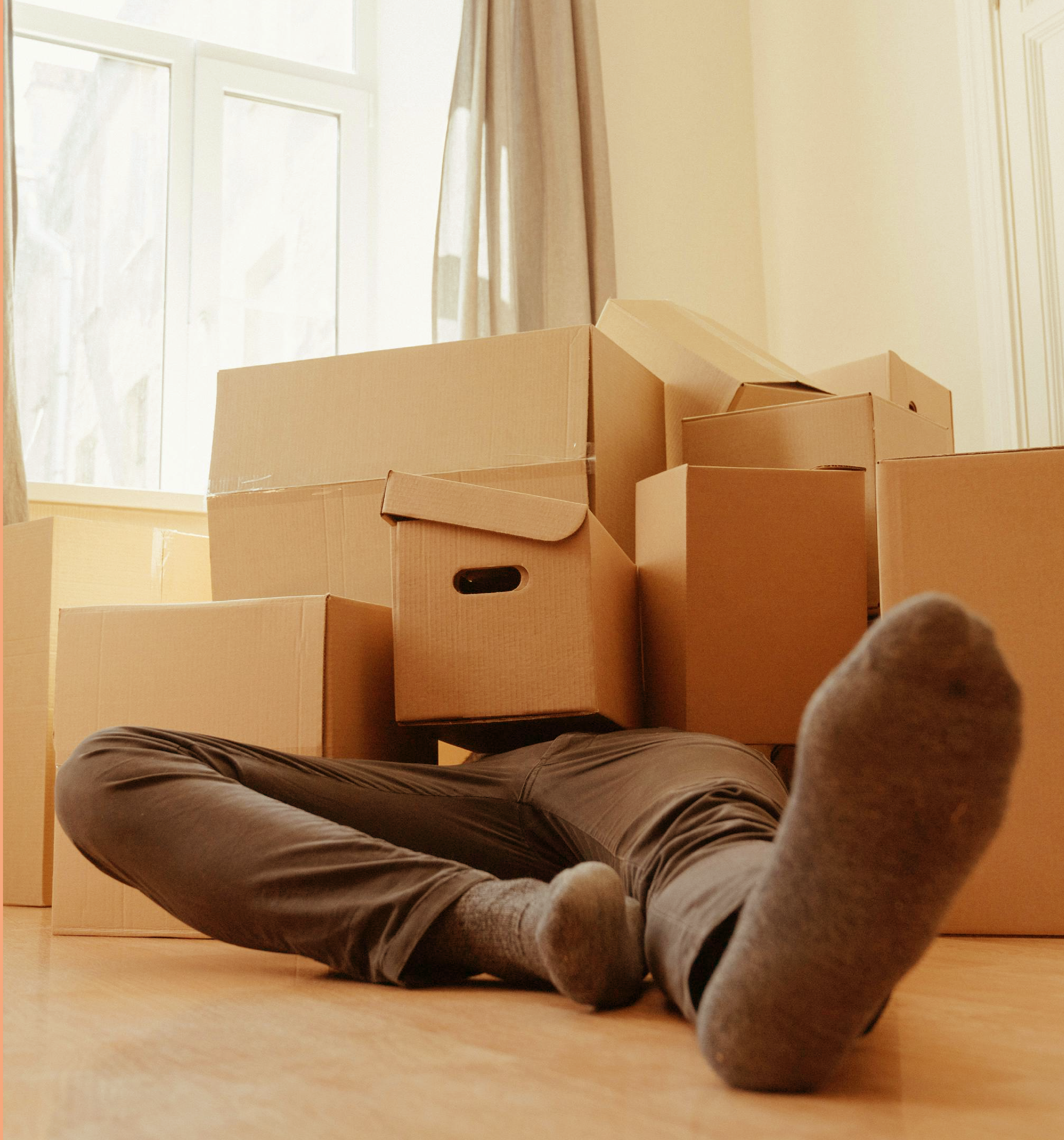 Person sitting on the floor with legs stretched out, wearing gray pants and socks, in a room with cardboard boxes and a window with blinds.