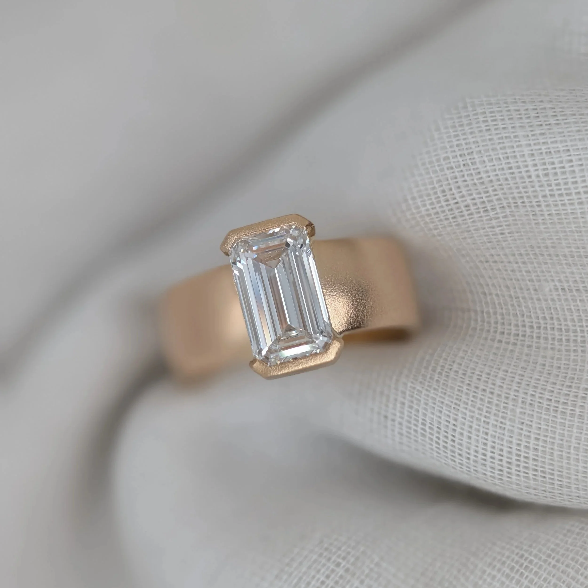 Photo of an emerald-cut diamond with top and bottom setting on a semi-flat gold band on a linen backdrop.