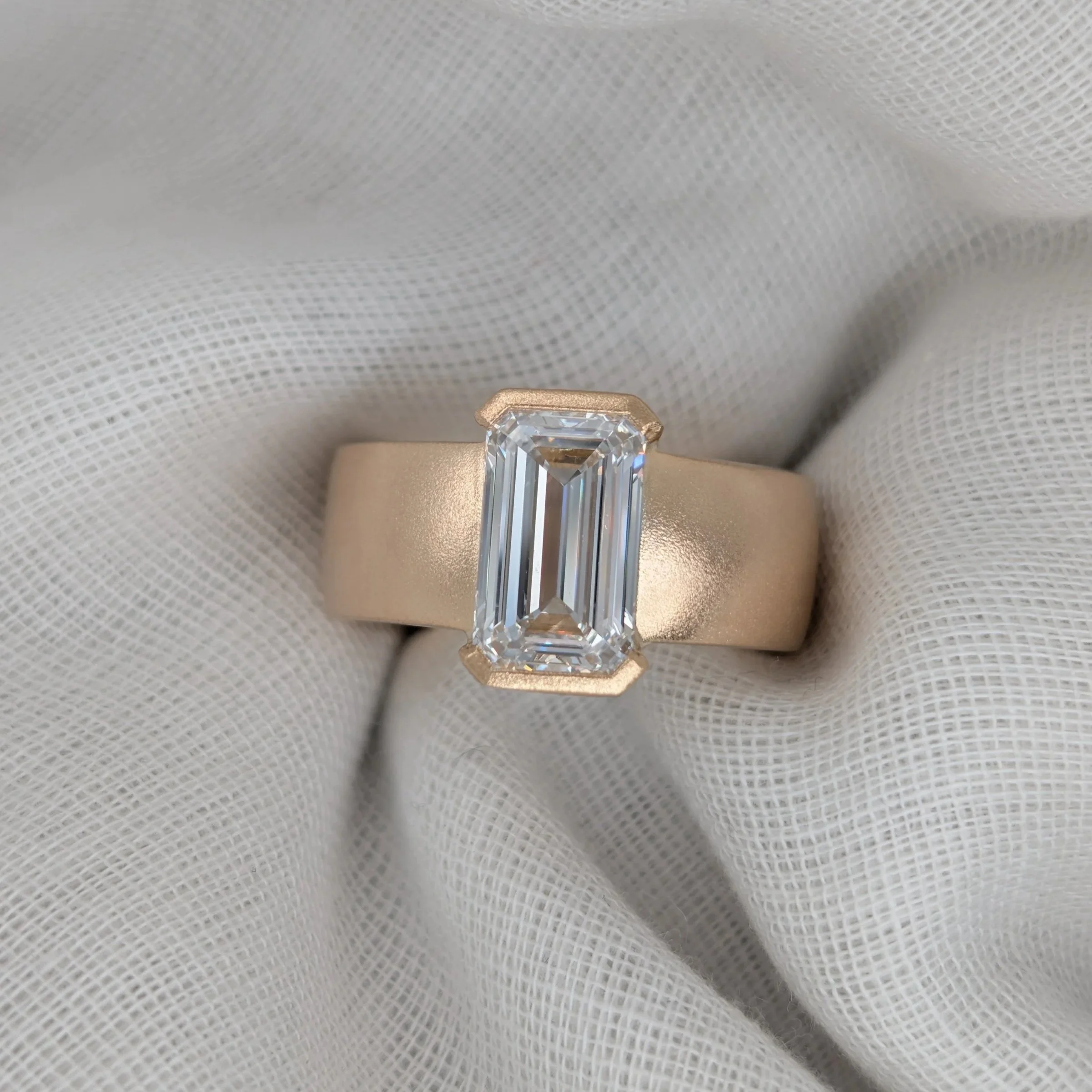 Photo of an emerald-cut diamond with top and bottom setting on a semi-flat gold band on a linen backdrop.