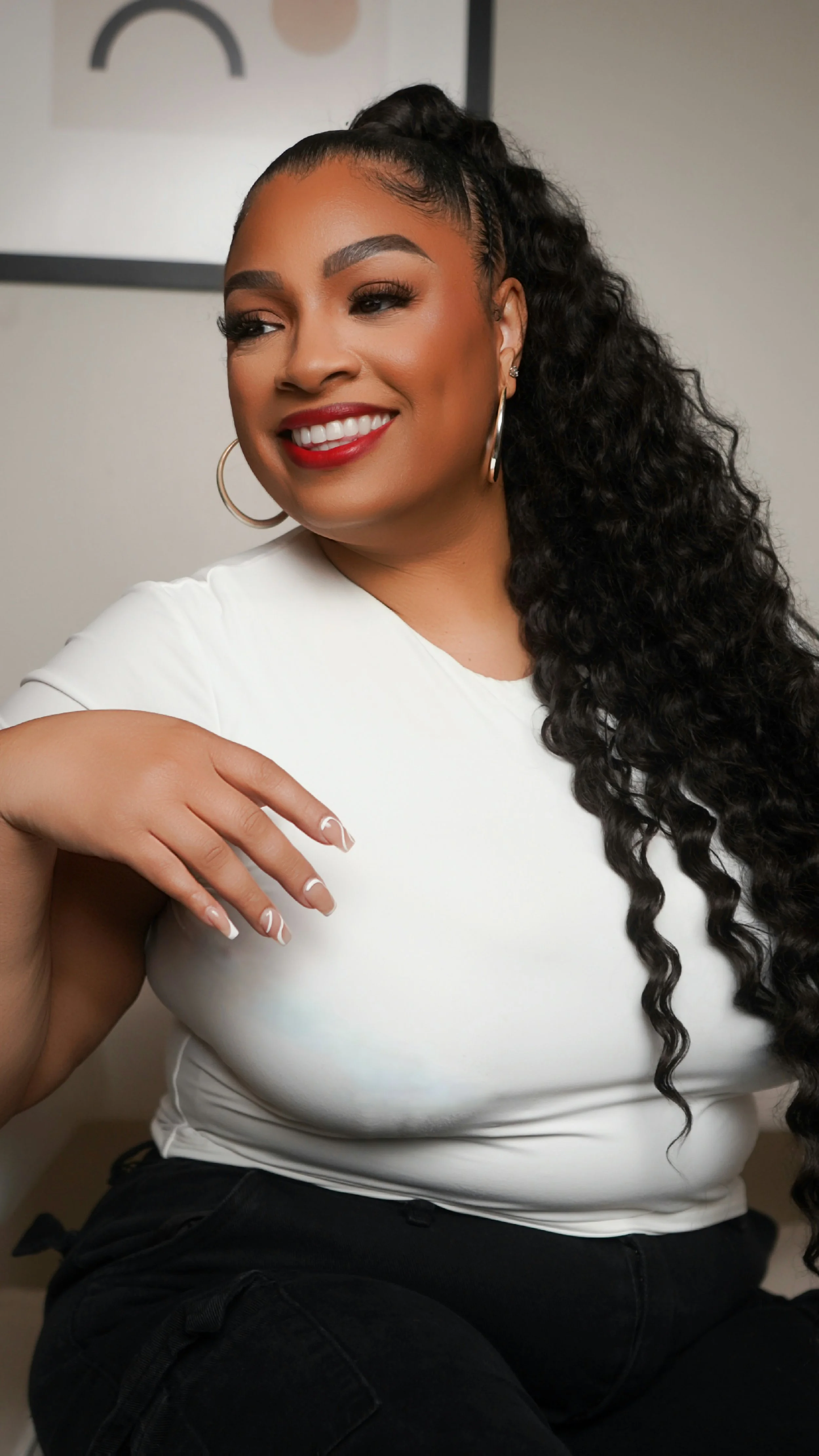 A woman with long, curly black hair, wearing hoop earrings, a white top, and black pants, smiling and looking to her right.