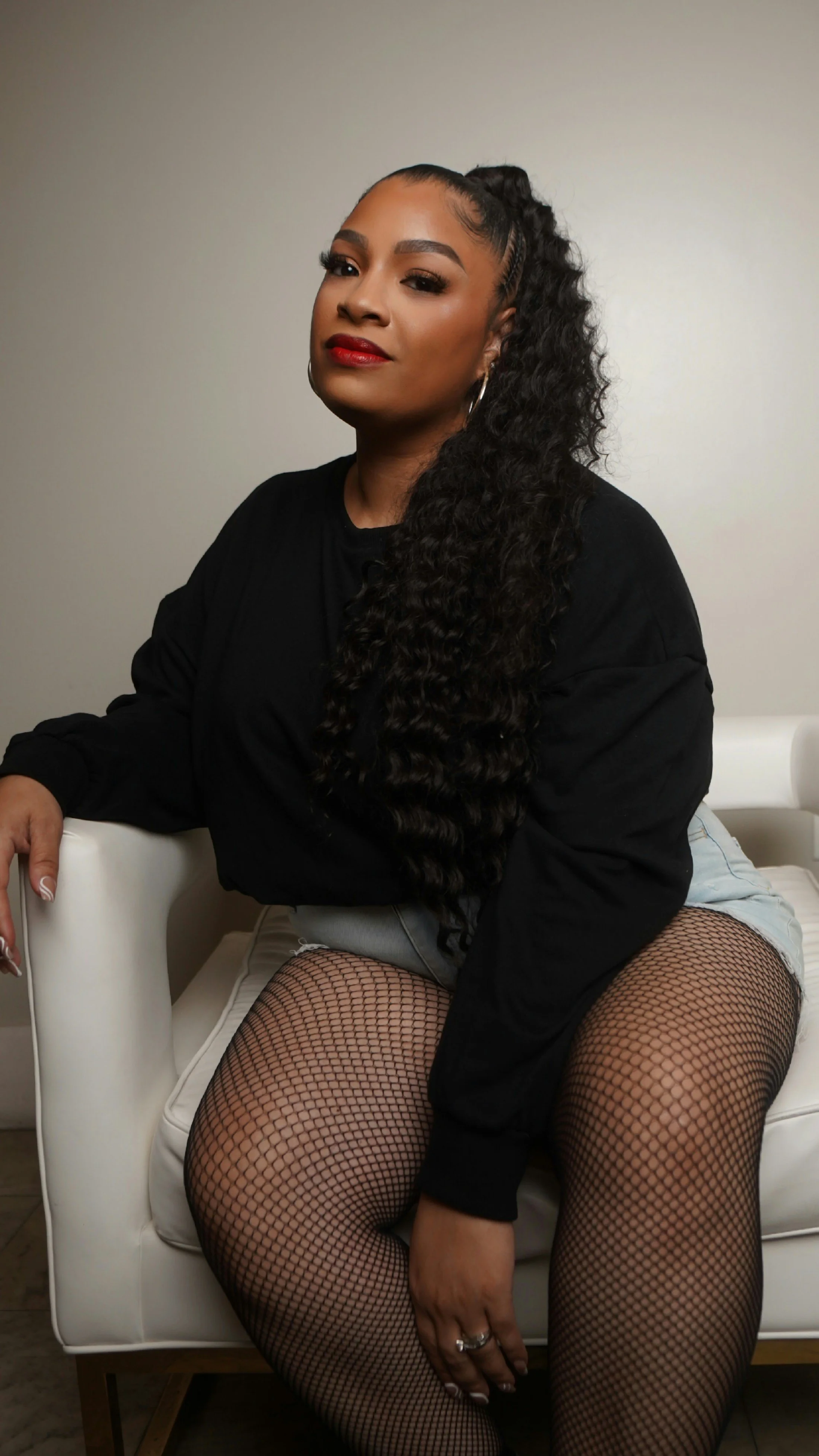 A woman with long, curly hair, wearing fishnet stockings, sitting on a white couch, dressed in a black top and denim shorts, with earrings and lipstick.