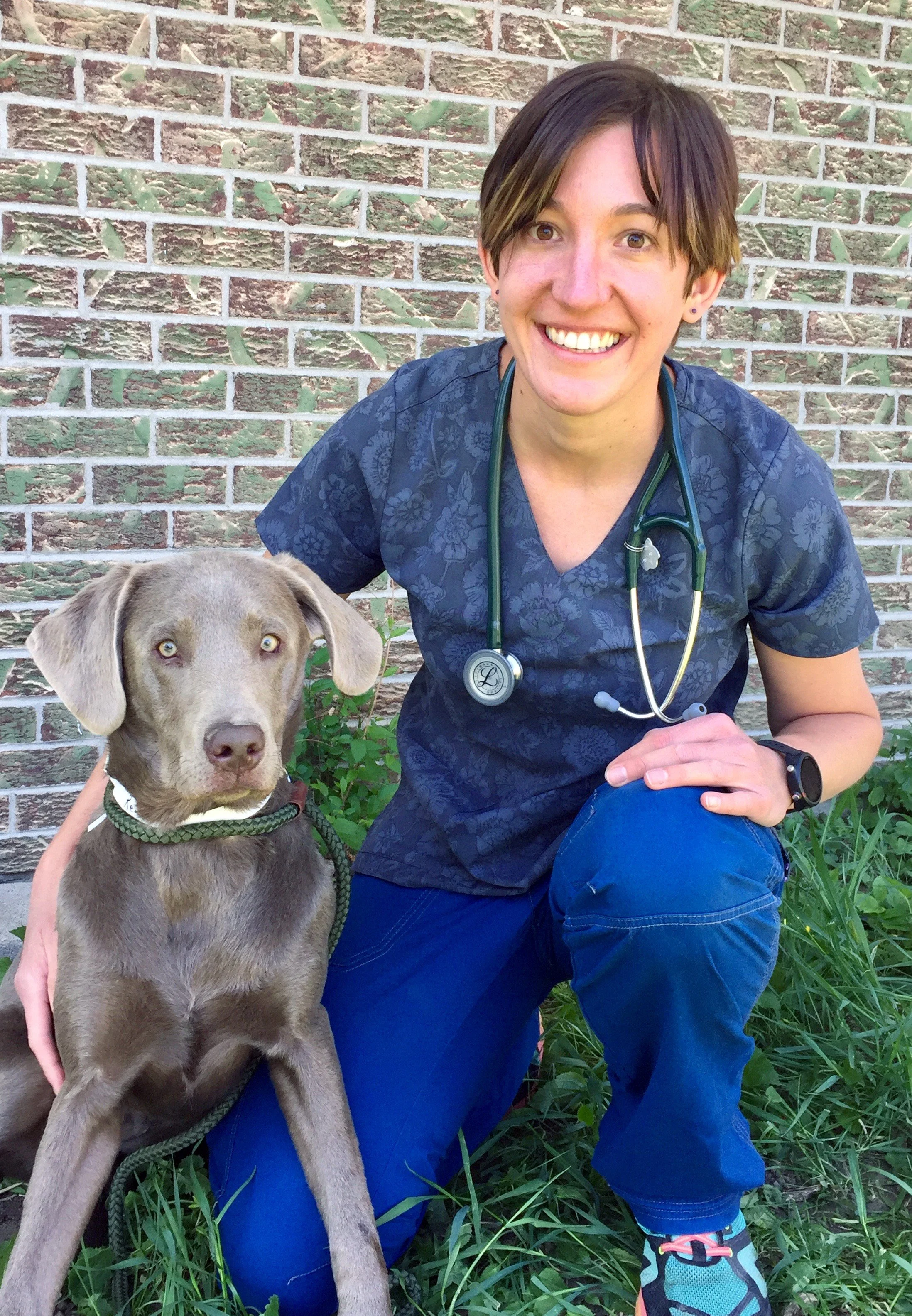Dr. Hanni Horner in blue scrubs with a stethoscope around her neck kneels next to a brown dog with floppy ears outside, in front of a brick wall and green grass.