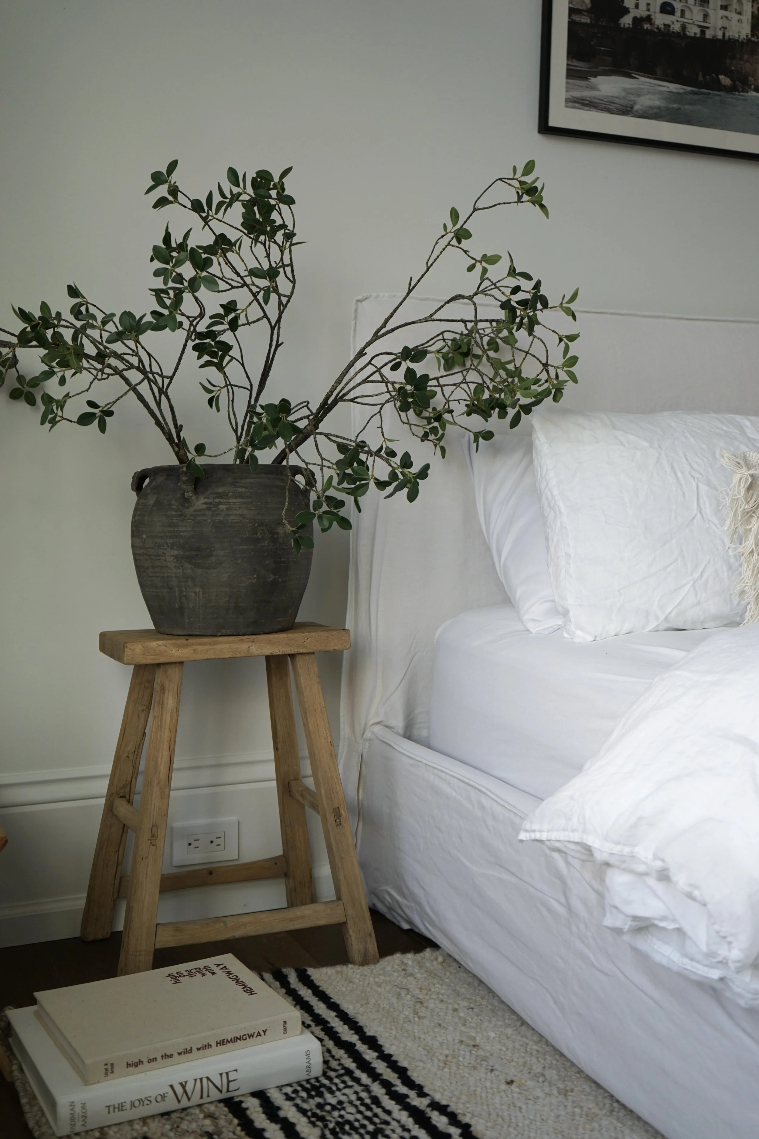 A cozy bedroom corner featuring a white bed with pillows and a slipcover, a rustic wooden stool with a large black pottery vase holding a leafy plant, and a stack of three books on a black and white striped rug.