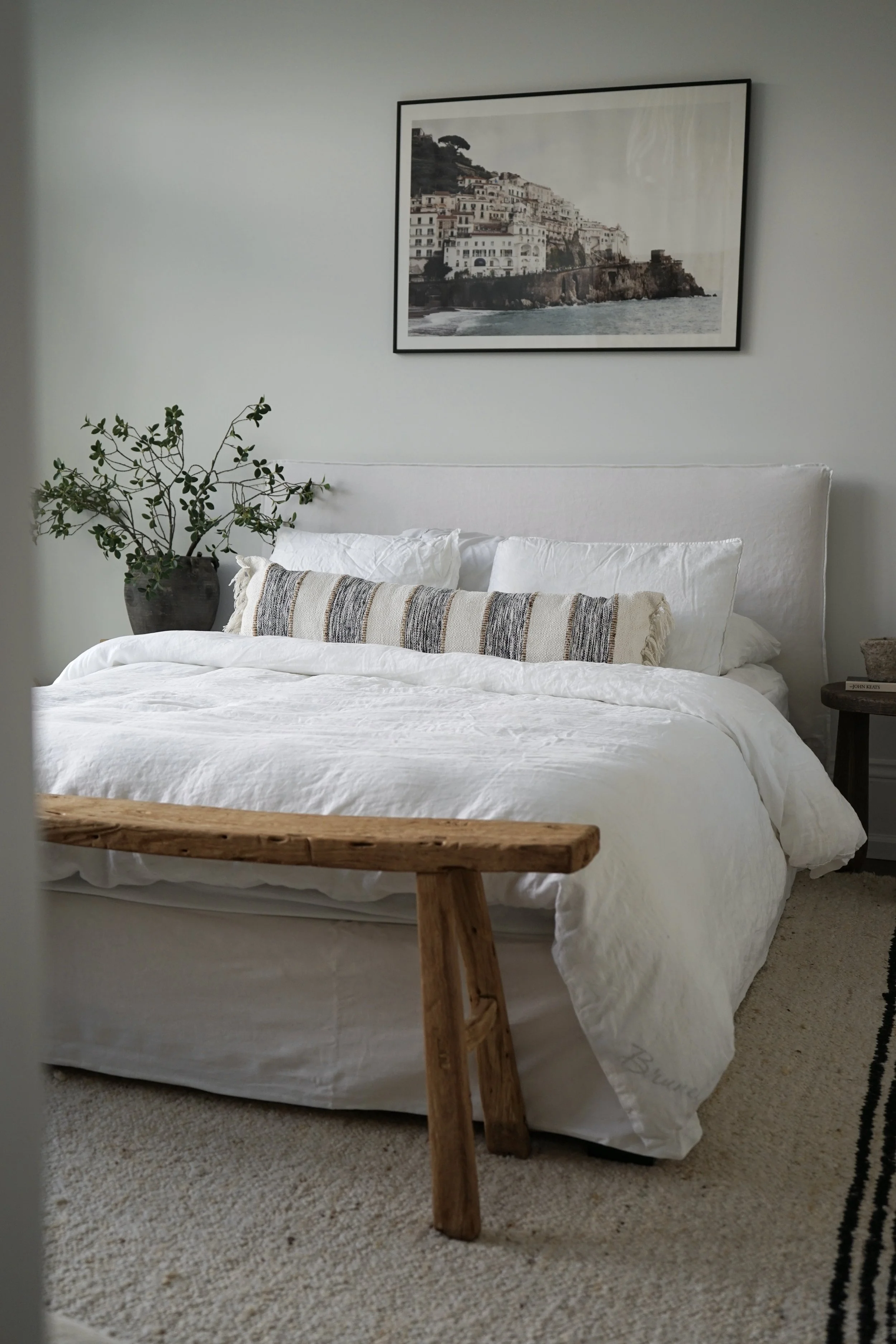 A cozy bedroom with a white bed with pillows and a striped decorative pillow, a wooden bench at the foot of the bed, a potted plant on a side table, wall art of a coastal town, and a beige rug.