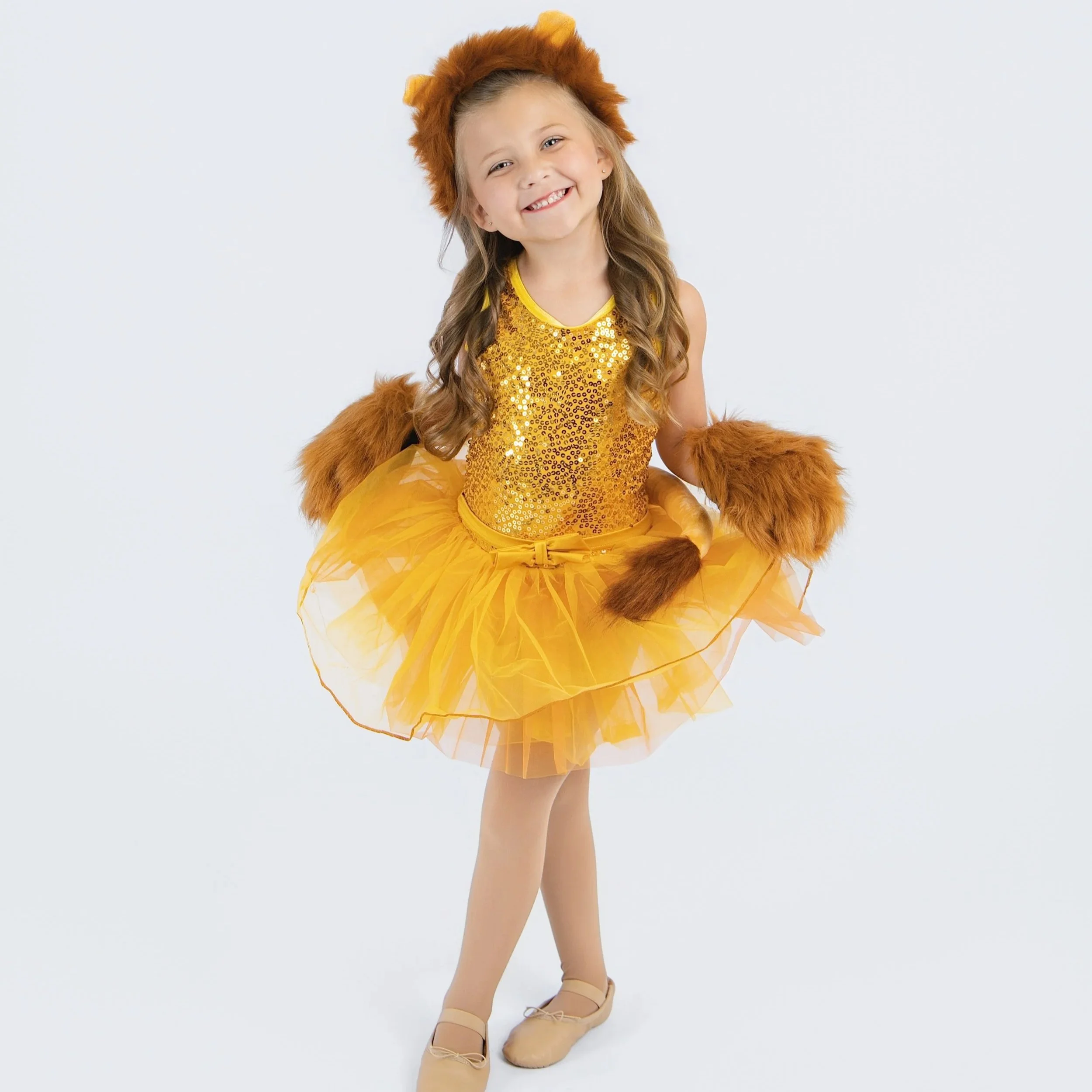 Young girl dressed in a yellow ballerina costume with a sequined top, yellow tutu, and a furry lion costume headpiece, standing with one leg slightly raised and smiling.