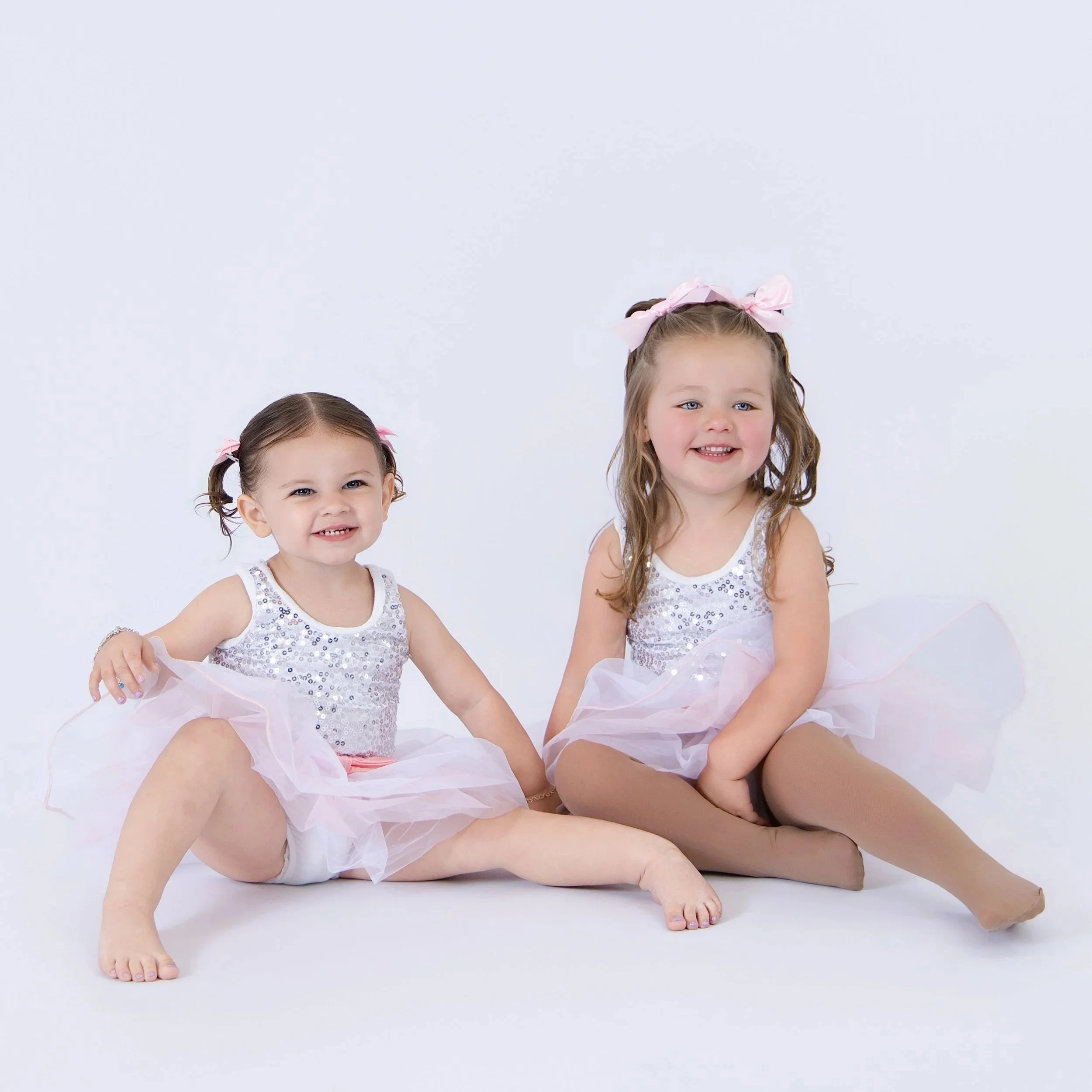 Two young girls with brown hair, wearing white and silver sequined dresses and pink hair bows, sitting together on the floor smiling at the camera against a light background.