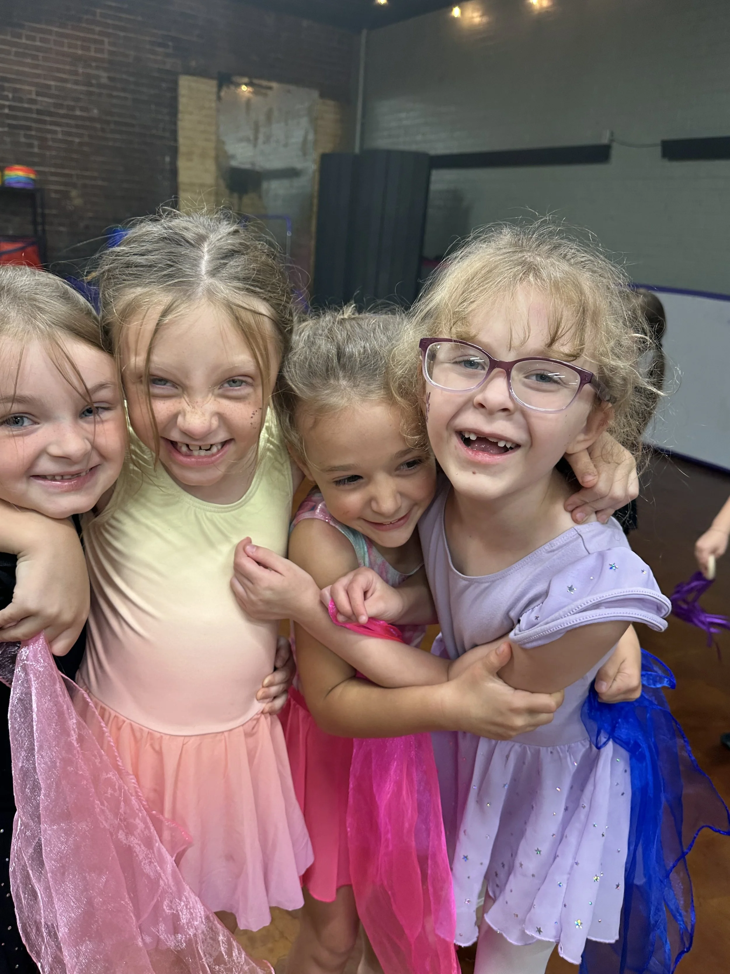 Four young girls in colorful fairy or princess costumes hugging and smiling in a room with brick walls.