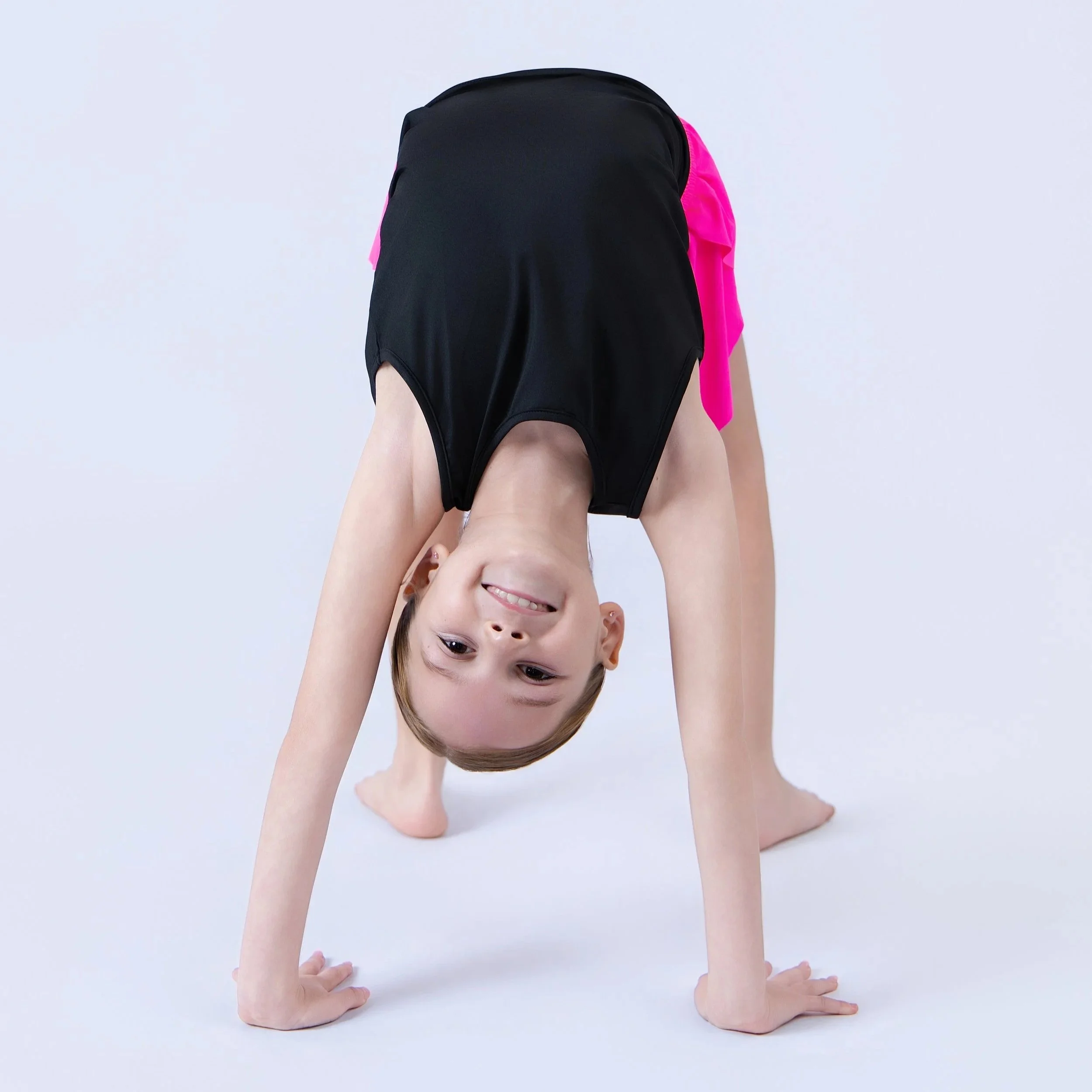 A young girl with brown hair and a big smile is upside down, supporting her body with her hands on a white surface, while her legs are bent at the knees, wearing black shorts and a bright pink top.