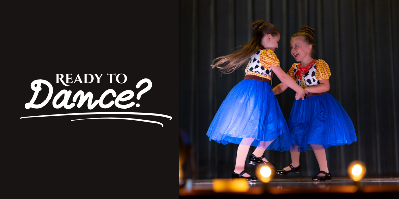 Two young girls in colorful costumes dance together on a stage with a dark curtain background, smiling and holding hands. The left side of the image has the text "Ready to Dance?" in white letters.