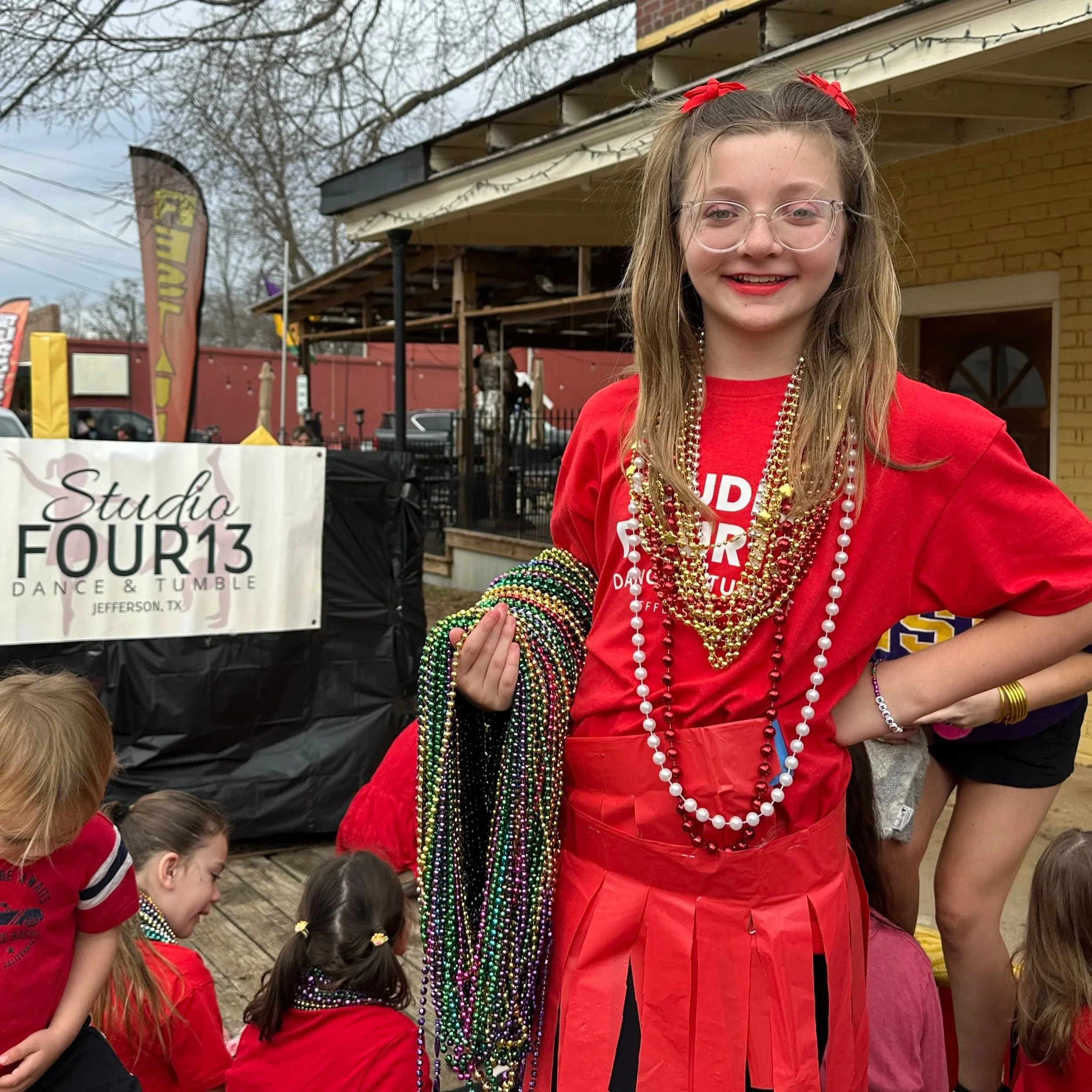 A young girl wearing a red shirt and red lipstick, decorated with red bows in her hair, is smiling and surrounded by children, wearing multiple colorful beads. The background shows an outdoor setting with a building, trees, and banners.