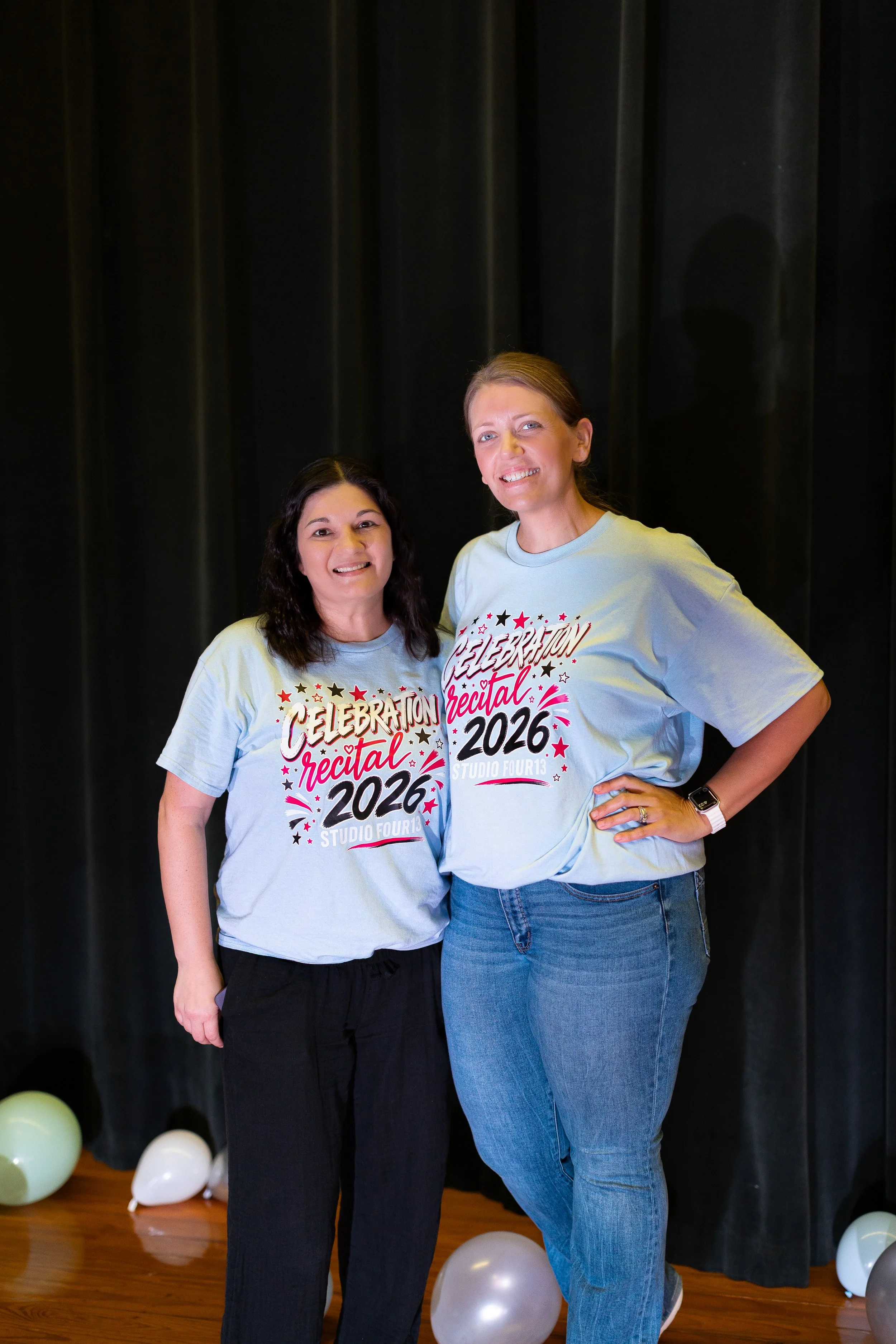 Two women standing in front of a black curtain, smiling, wearing matching light grey shirts with colorful text and stars that read 'Celebration Recital 2026 Studio Four13'. They are surrounded by balloons in various colors on the floor.