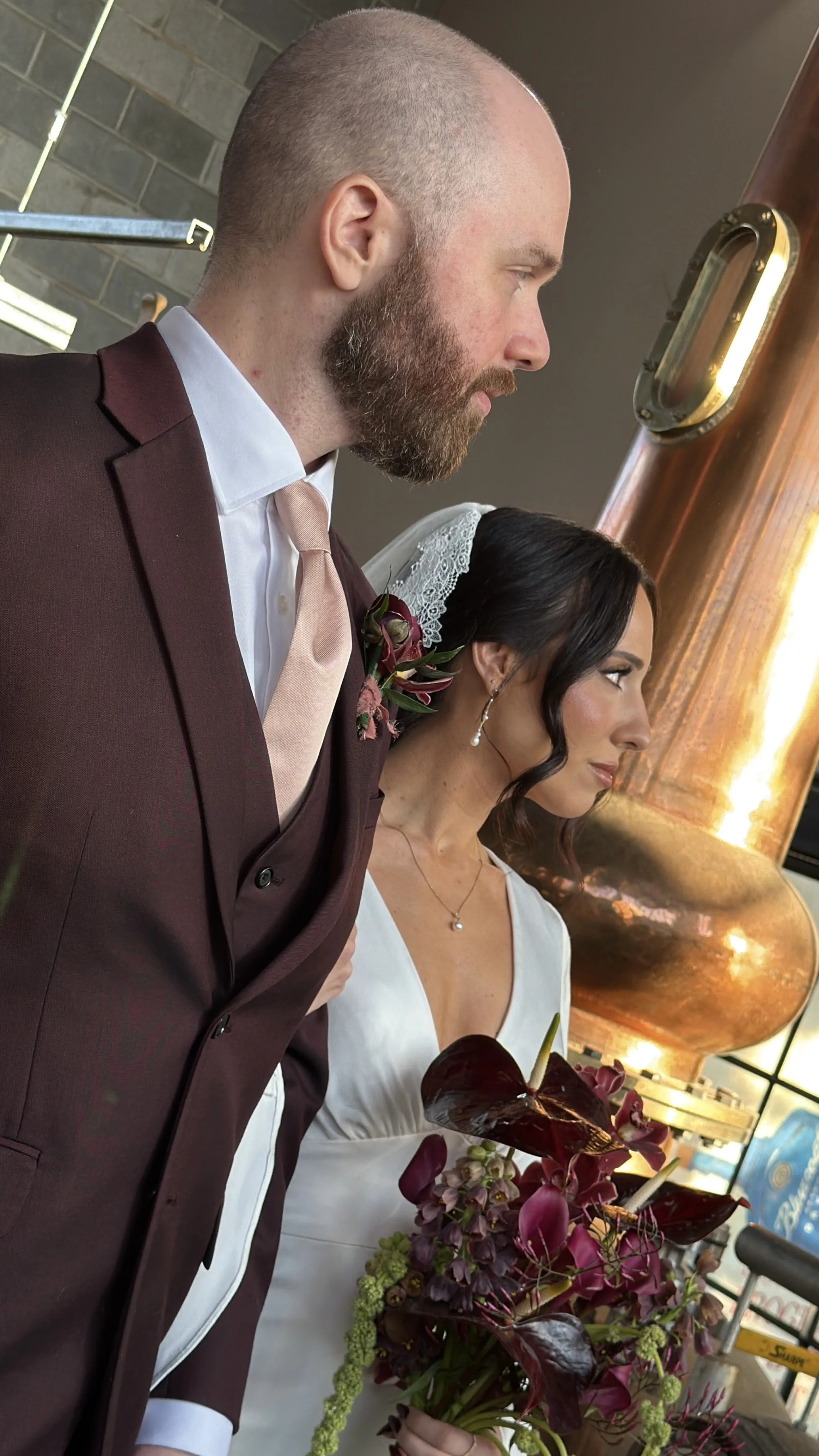 A bride and groom standing side by side, with the groom in a burgundy suit and the bride in a white dress, holding a floral bouquet, at a wedding ceremony.
