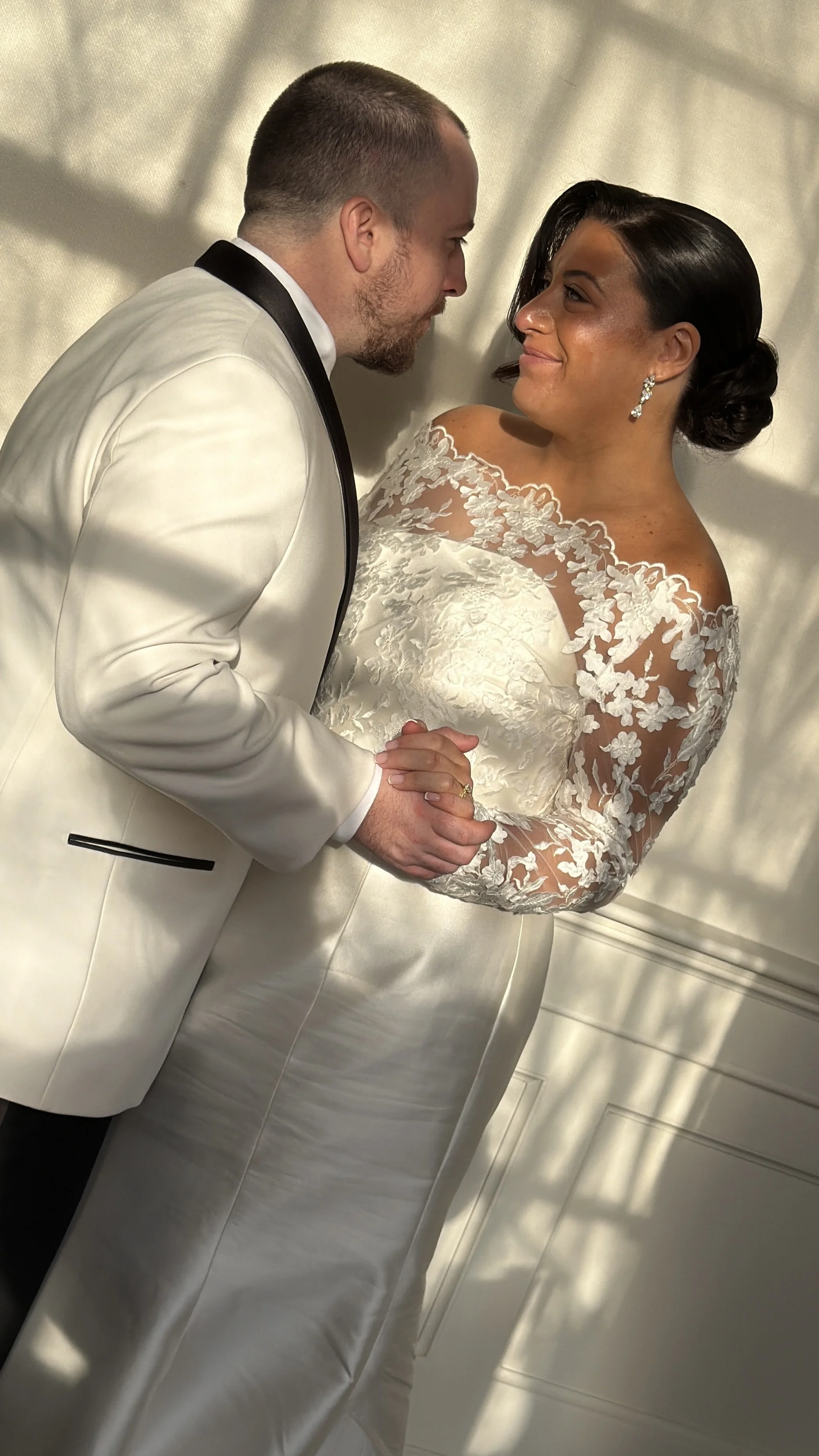 A man in a white tuxedo and a woman in a white lace wedding dress share a tender moment, holding hands and leaning in close for a kiss.