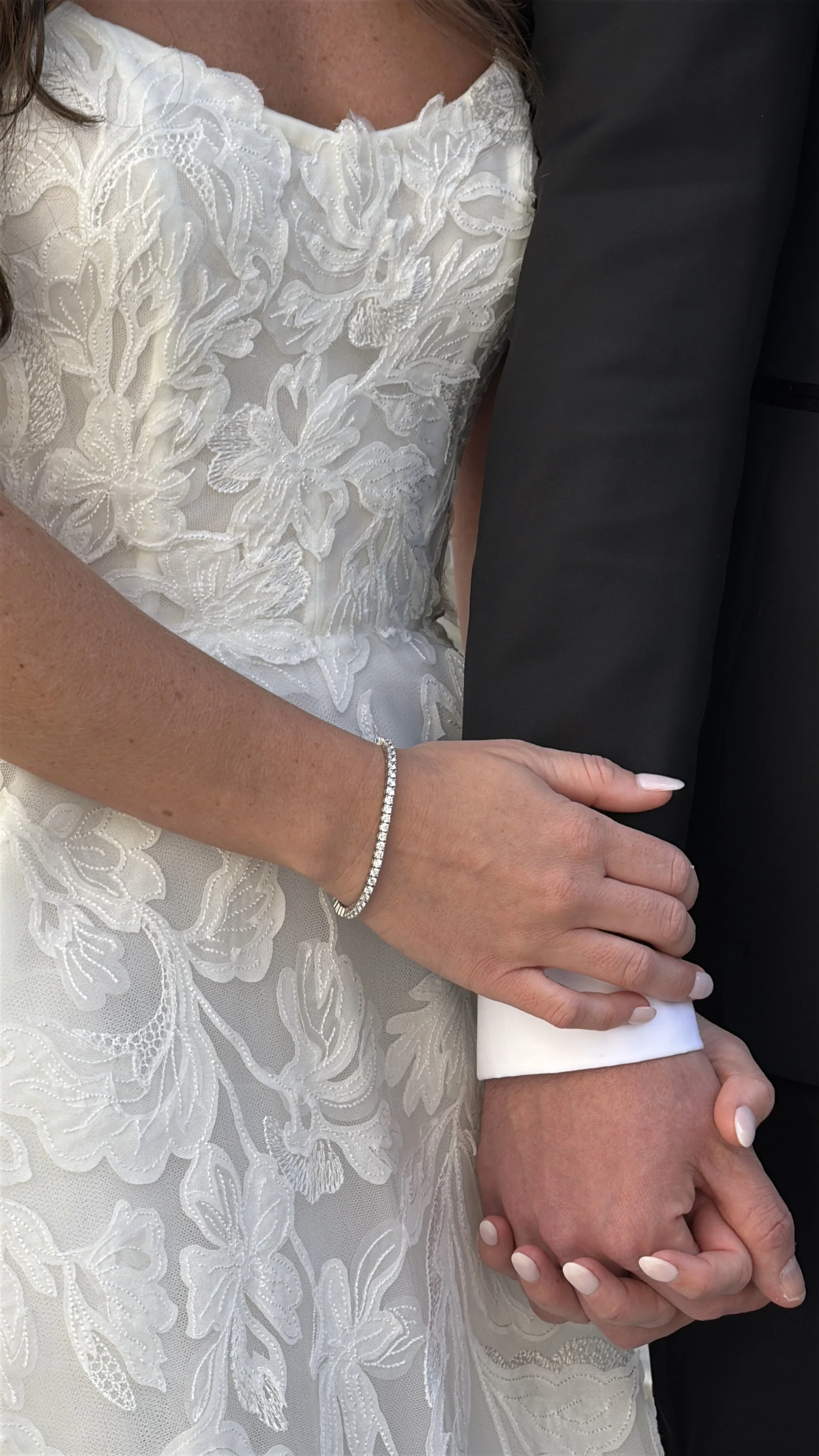 Close-up of a bride in a white lace wedding dress holding hands with a person in a dark suit, with a diamond bracelet on her wrist.