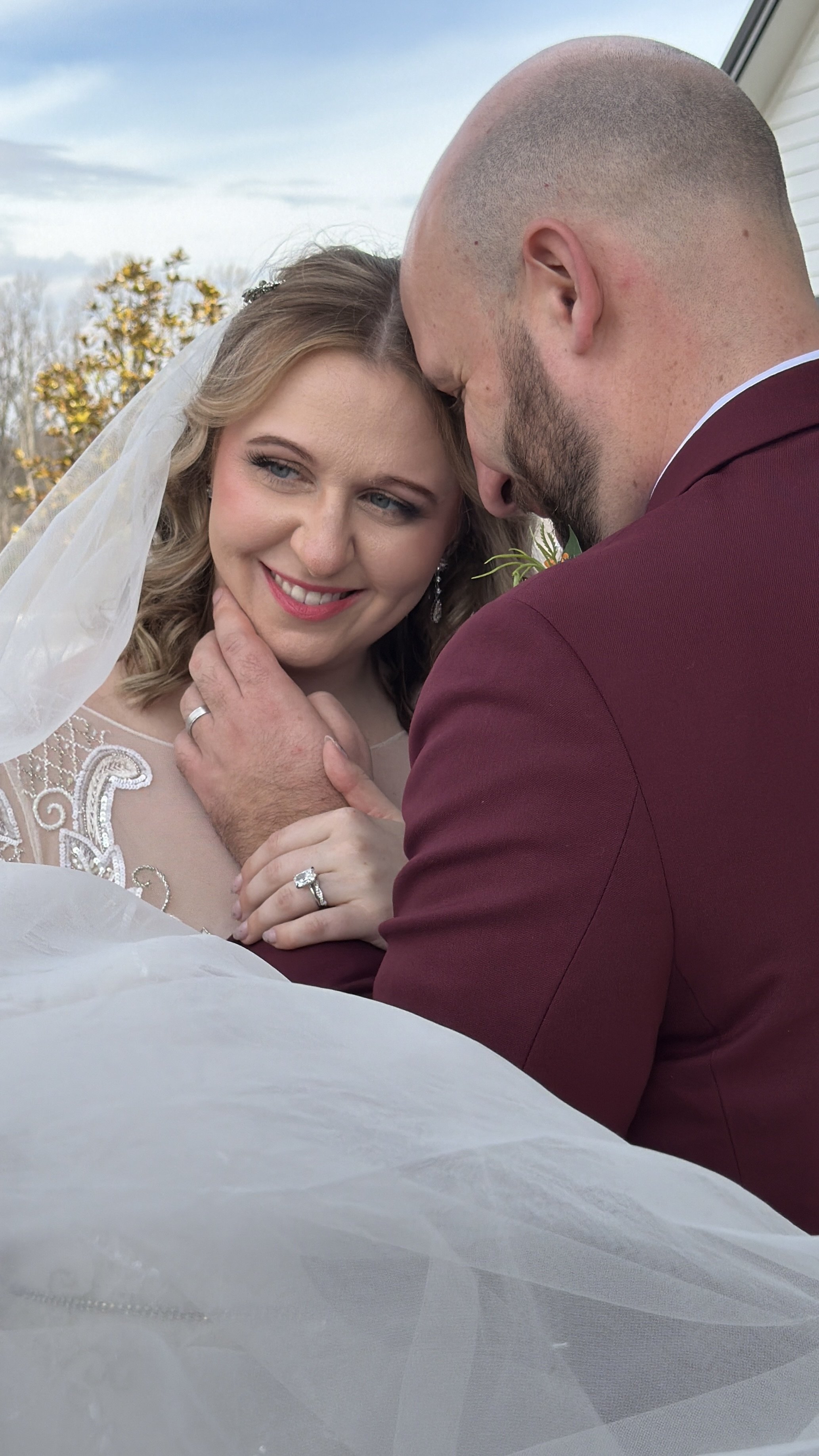 A bride and groom share a loving moment, with the bride smiling and the groom gently holding her face, outdoors with trees and a cloudy sky in the background.
