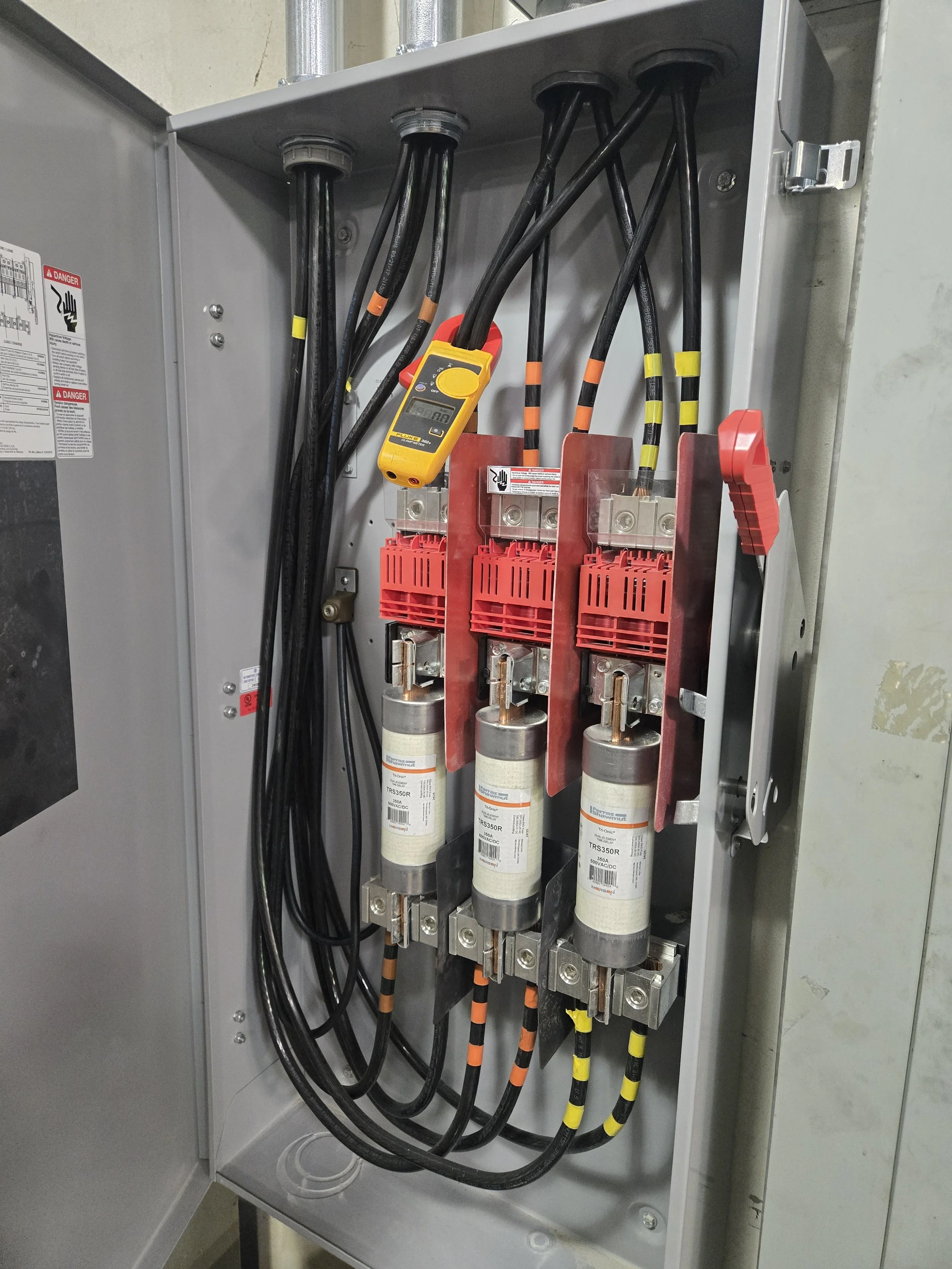 Electrical panel with three large circuit breakers, in-line yellow and black multimeter, and multiple black cables with colored tape for identification.