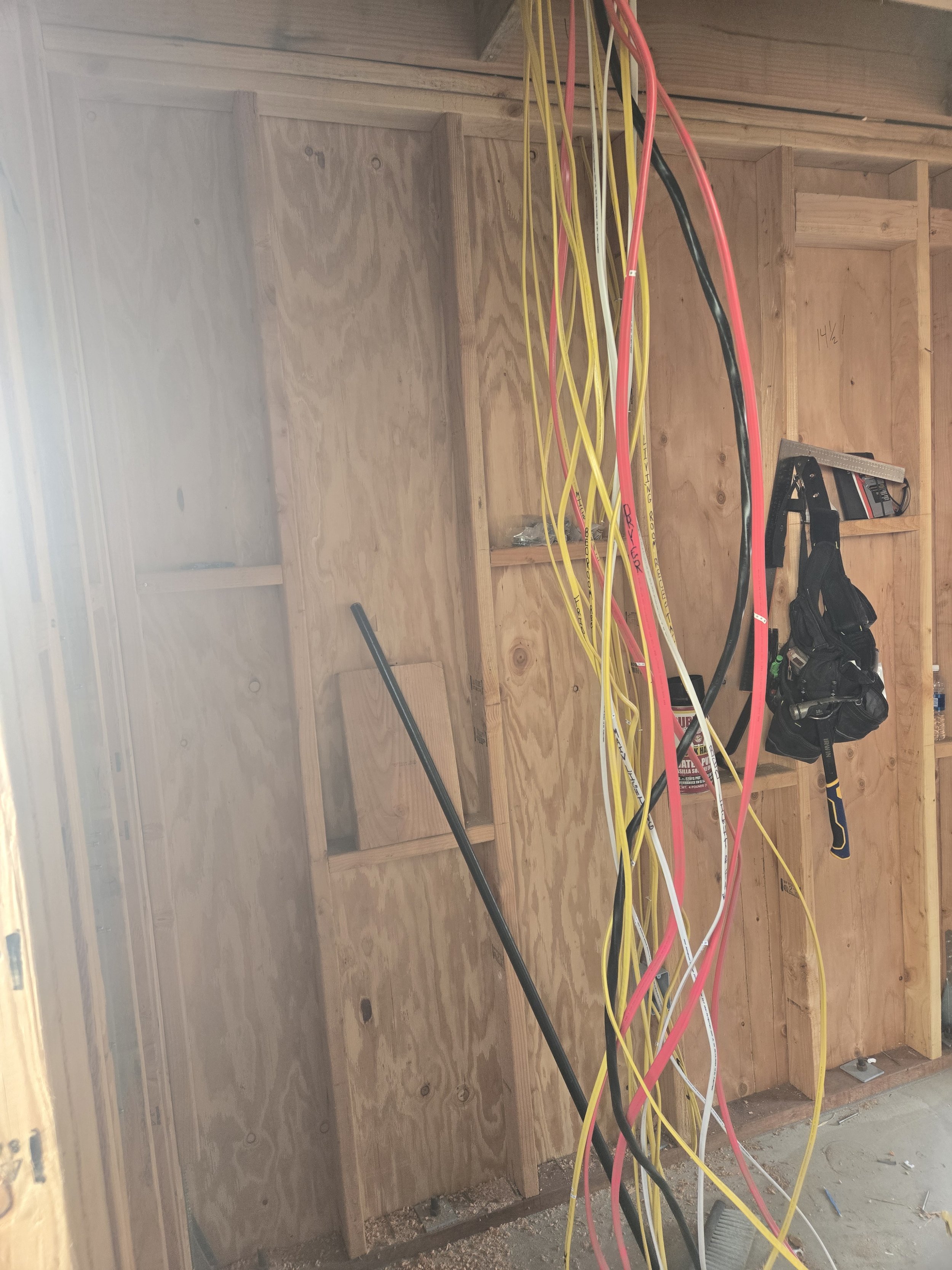 Interior of a building under construction with electrical wires hanging from the ceiling, a wooden wall framing, a black tool belt, and a saw in the background.