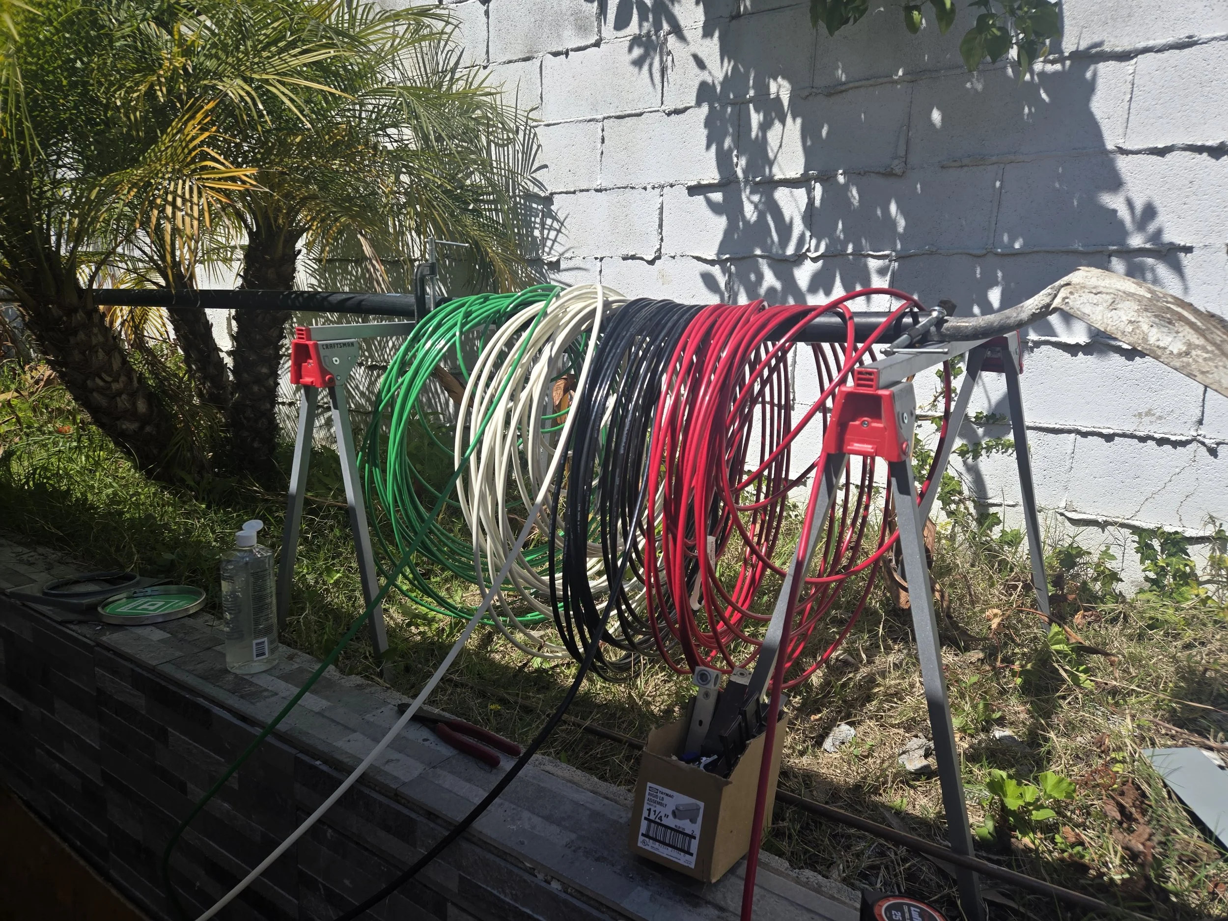 Multiple colored extension cords hanging on a stand outdoors near a white brick wall and palm trees.
