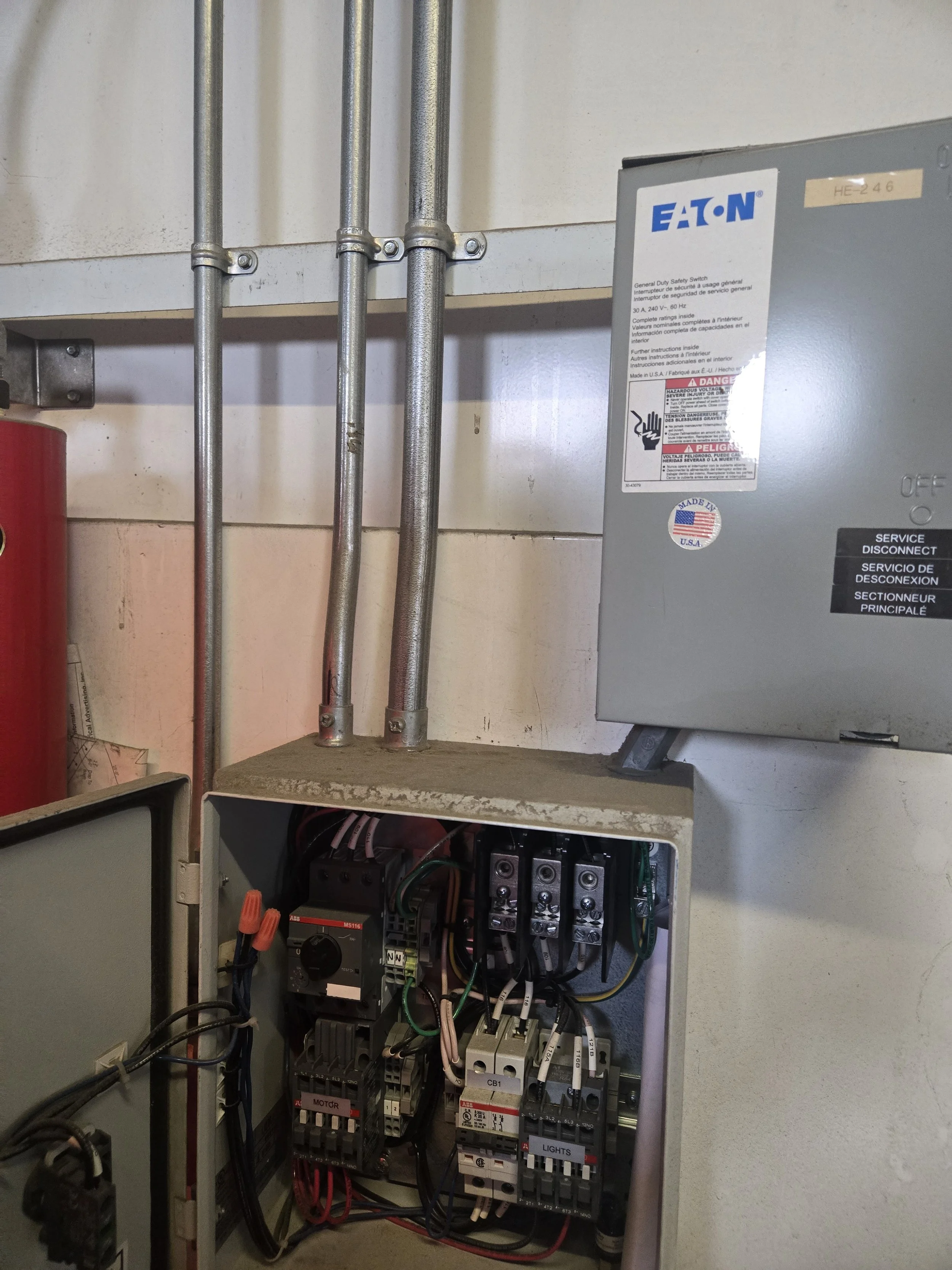 Electrical panel with wires and switches next to an Eaton safety switch box on a beige wall.